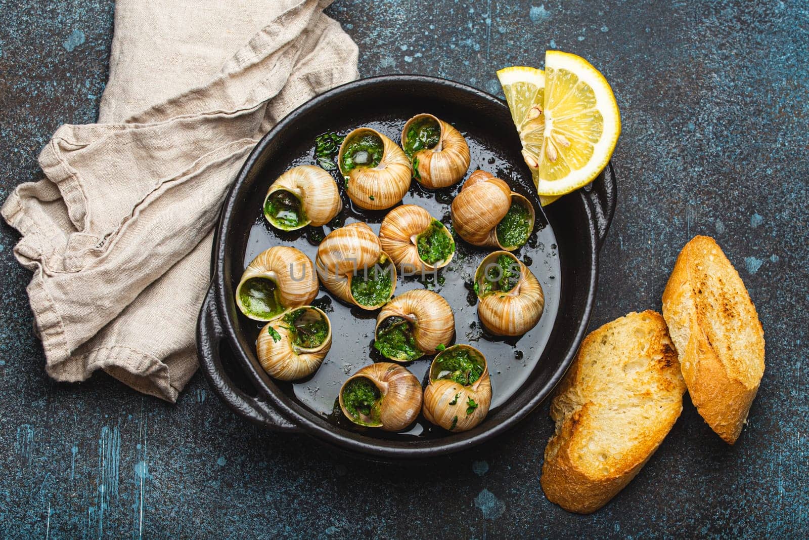 Escargots de Bourgogne Snails with Garlic Butter and Parsley in black cast iron pan with Lemon and Toasted Baguette Slices on rustic stone background top view, traditional French Delicacy .