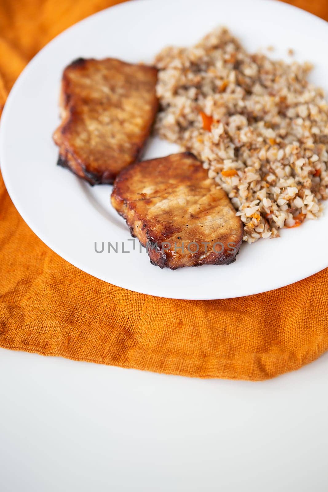 Delicious and appetizing pork tenderloin steak, grilled with buckwheat and vegetables, is on the table along with an orange napkin. by sfinks