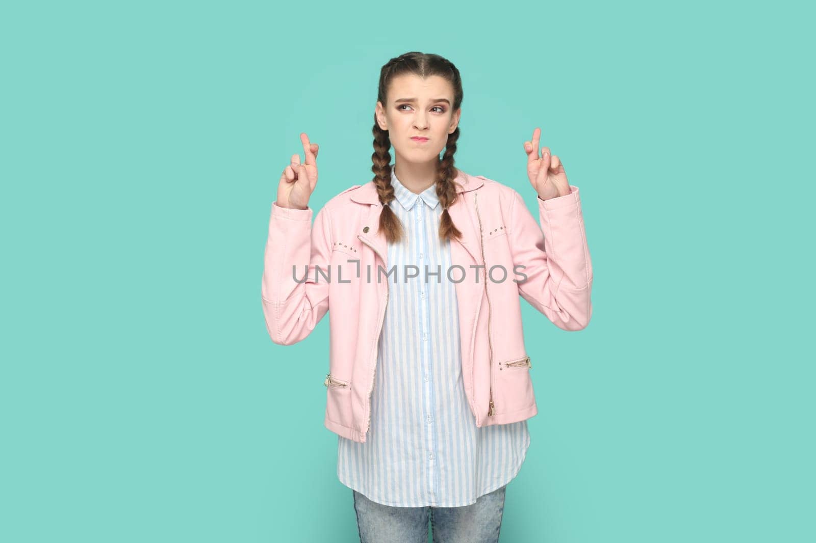 Portrait of beautiful teenager girl with braids wearing pink jacket standing with crossed fingers, making wish, desire, looking away. Indoor studio shot isolated on green background.