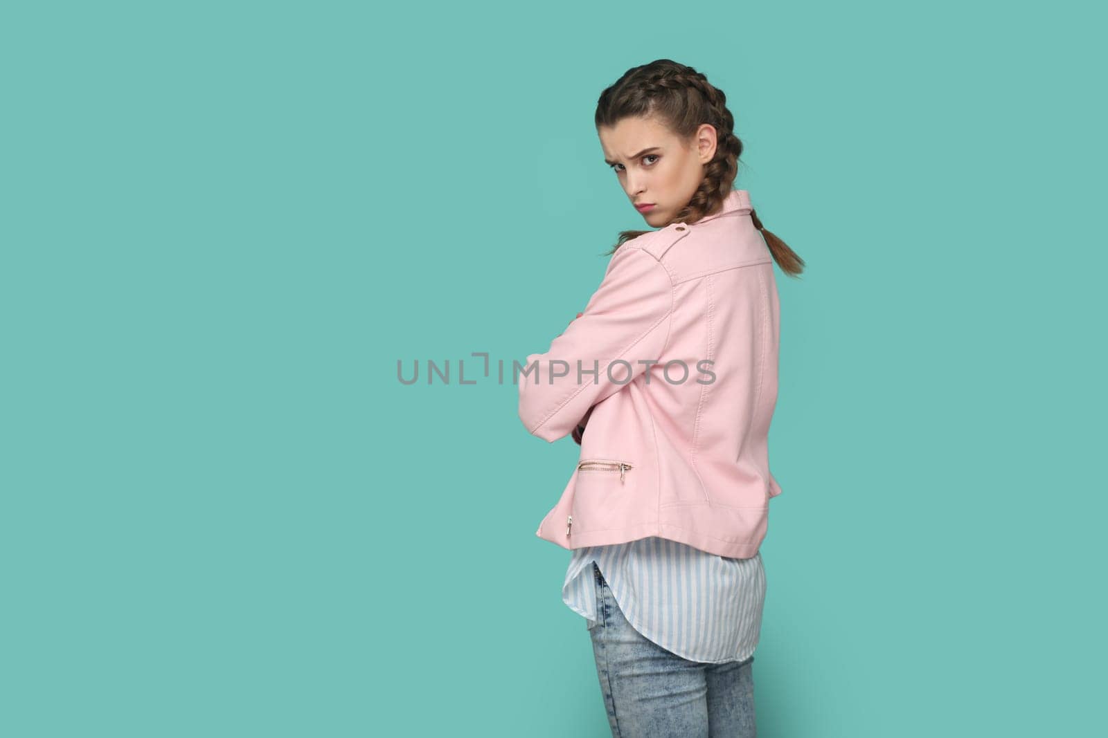 Portrait of upset teenager girl with braids wearing pink jacket turning to camera with sad look, looking at camera, expressing sorrow and sadness. Indoor studio shot isolated on green background.