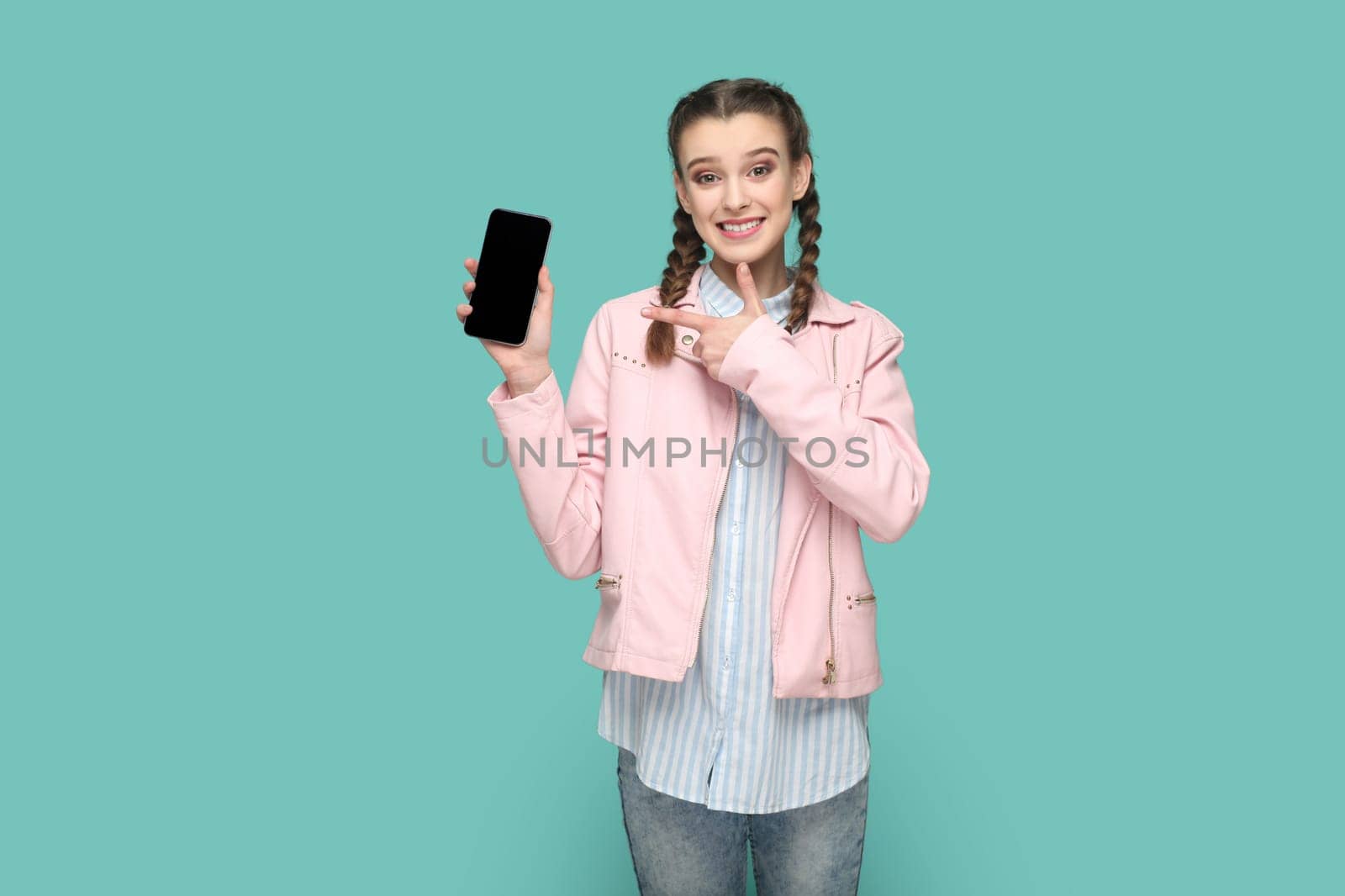 Portrait of cheerful joyful satisfied teenager girl with braids wearing pink jacket showing pointing at smart phone with empty display. Indoor studio shot isolated on green background.