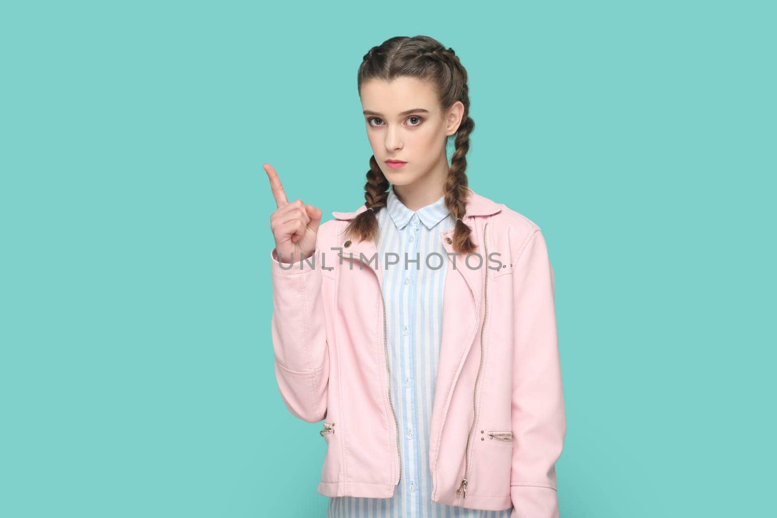 Portrait of serious bossy teenager girl with braids wearing pink jacket standing with raised finger, warning her classmates about danger. Indoor studio shot isolated on green background.