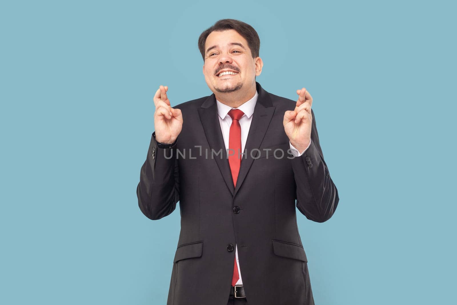 Portrait of smiling satisfied man with mustache standing keeps finger crossed, making wish, praying for good luck, wearing black suit with red tie. Indoor studio shot isolated on light blue background