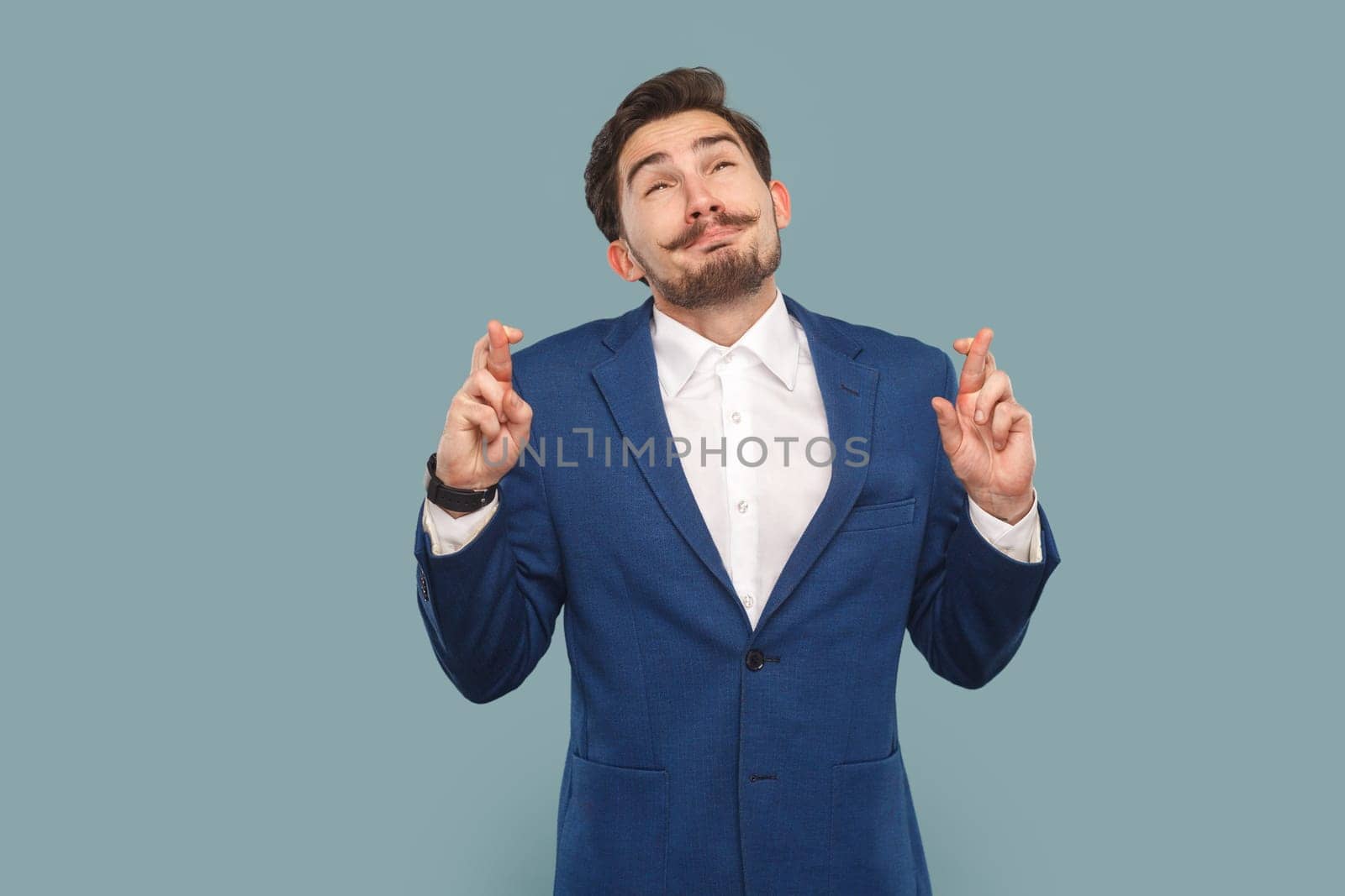 Portrait of hopeful handsome man with mustache standing with crossed fingers, hopes for better and good luck, wearing white shirt and jacket. Indoor studio shot isolated on light blue background.