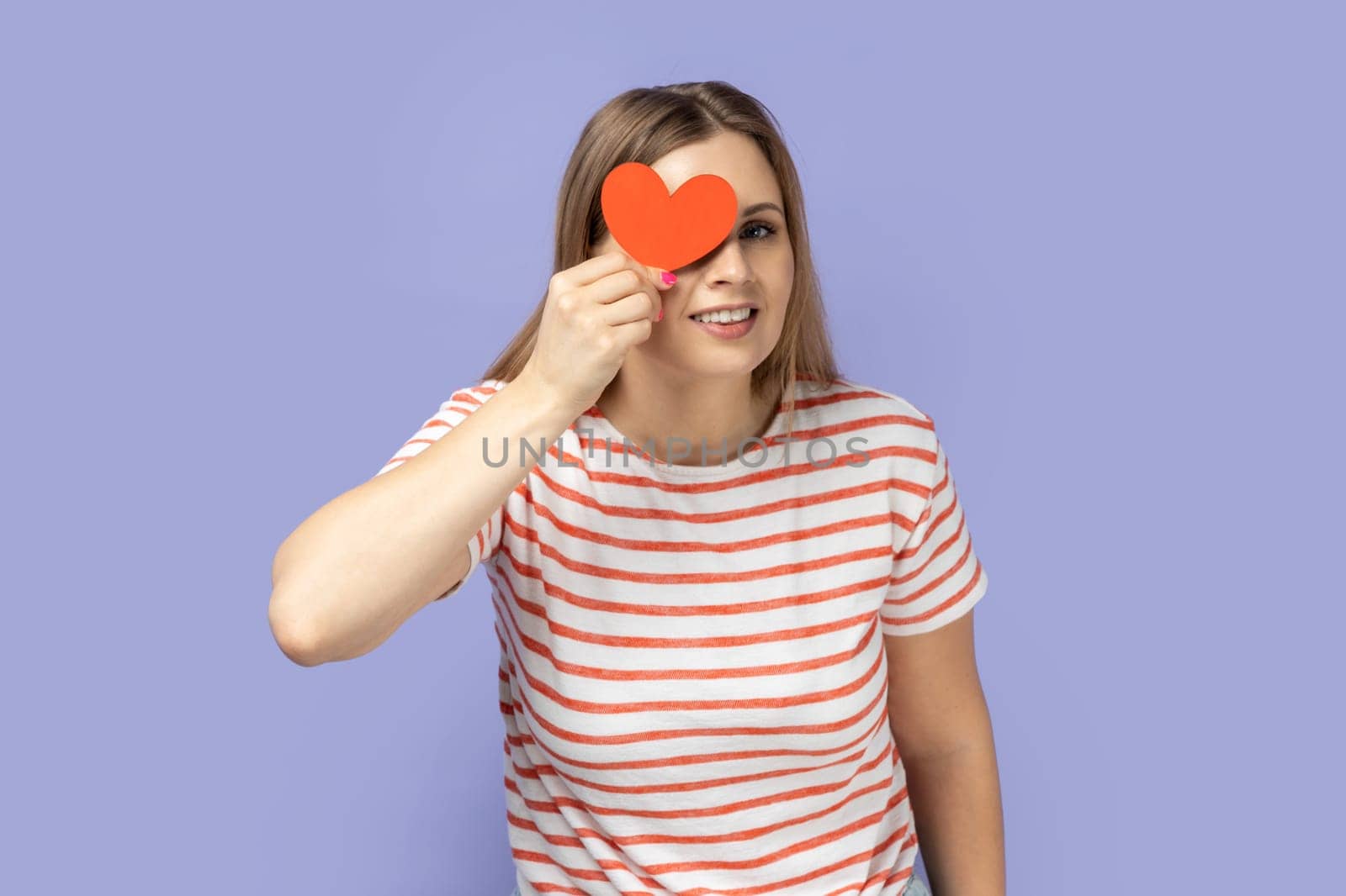 Positive funny woman holding little red heart and covering her eye behind love symbol. by Khosro1