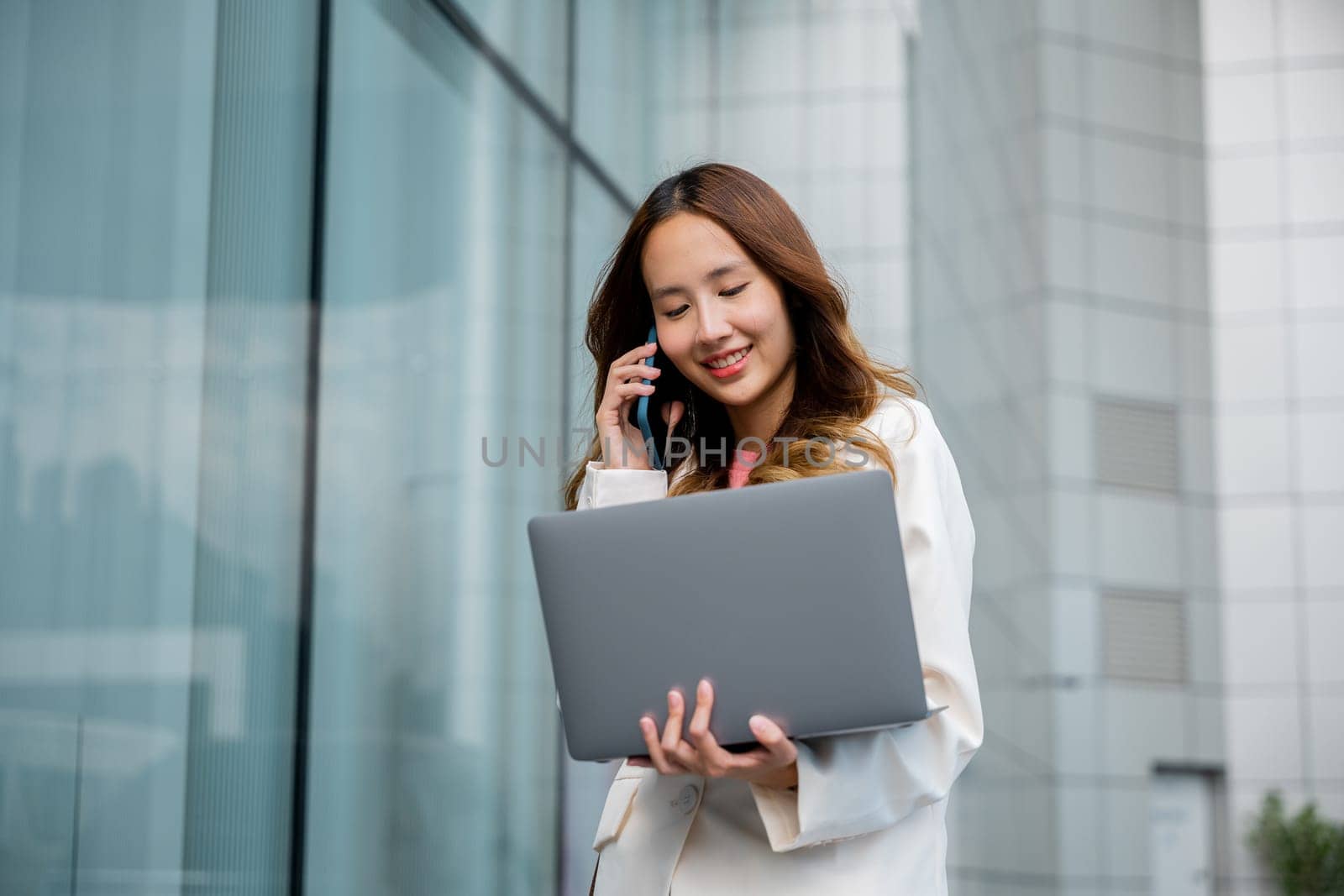 Portrait beautiful business woman smiling holding computer discussing issues on smart mobile phone in city, Asian businesswoman working on laptop and talking cell phone at front building near office