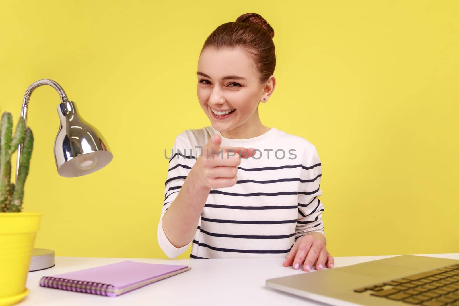 Delighted woman blogger pointing at you, looking at camera with toothy smile, POV, point of view of photo, while sitting at workplace. Indoor studio studio shot isolated on yellow background.