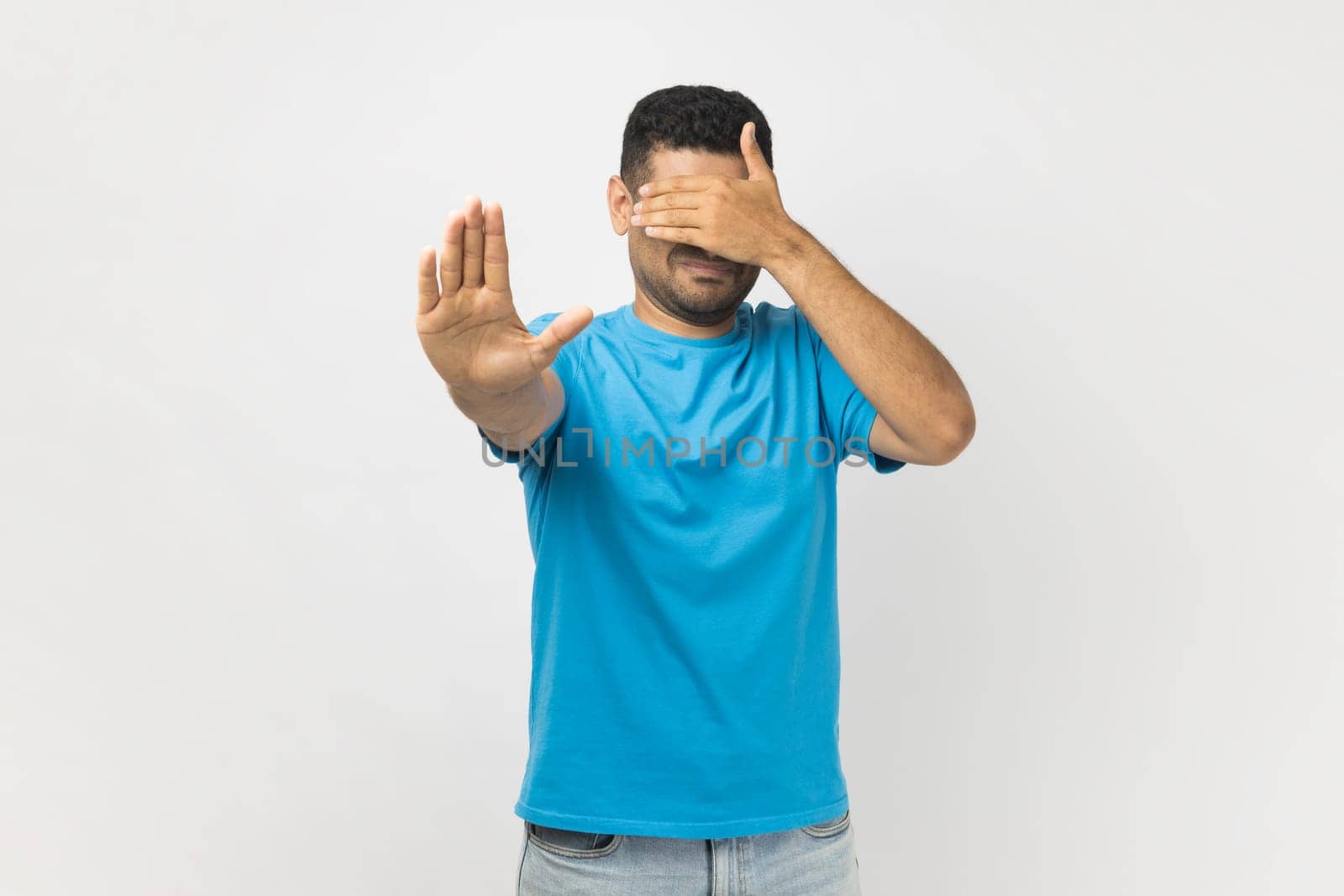 Portrait of young adult unshaven man wearing blue T- shirt standing covering his face with hand, hiding his face, making stop gesture. Indoor studio shot isolated on gray background.
