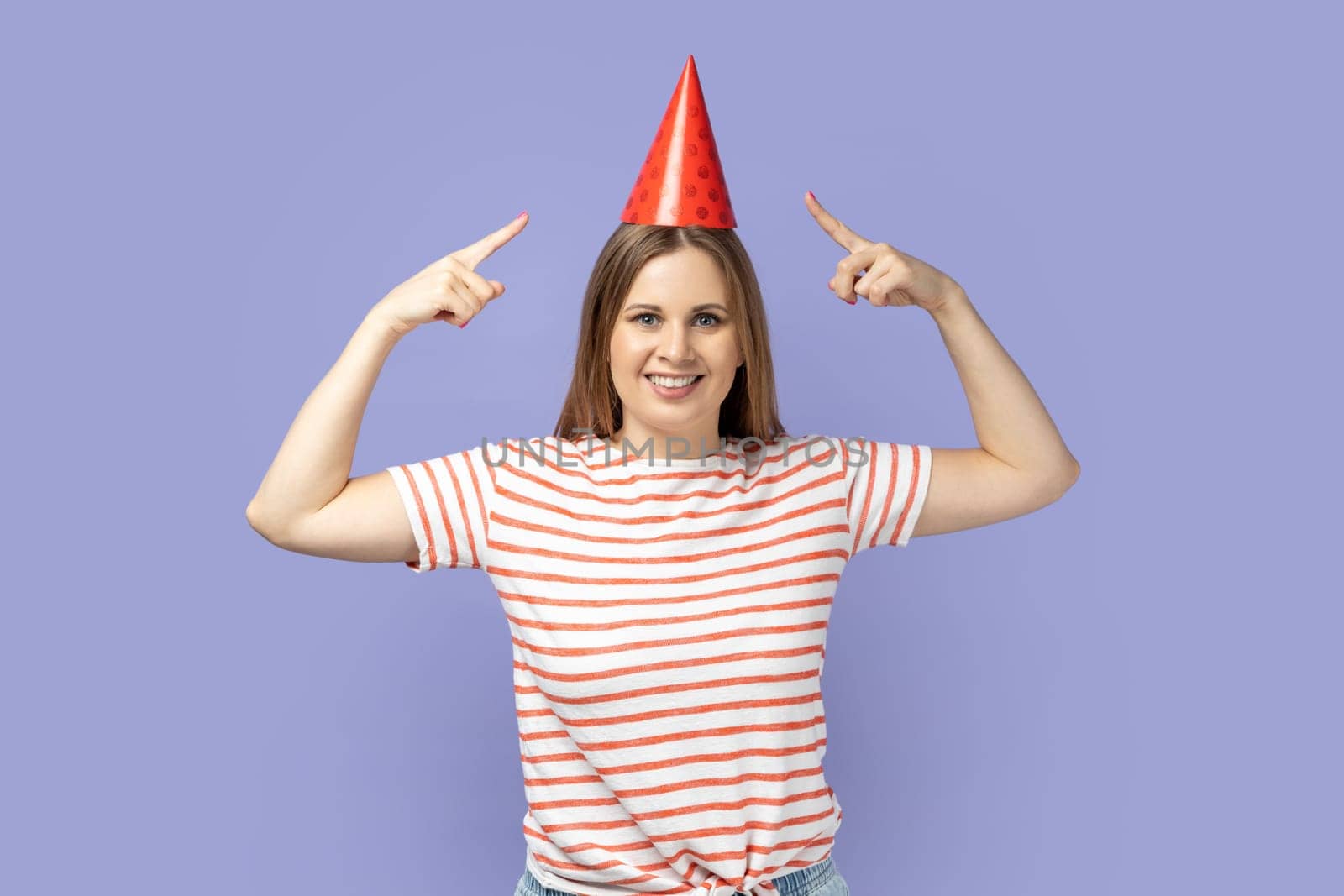 Woman pointing to funny party cone on head and smiling to camera, celebrating career growth. by Khosro1