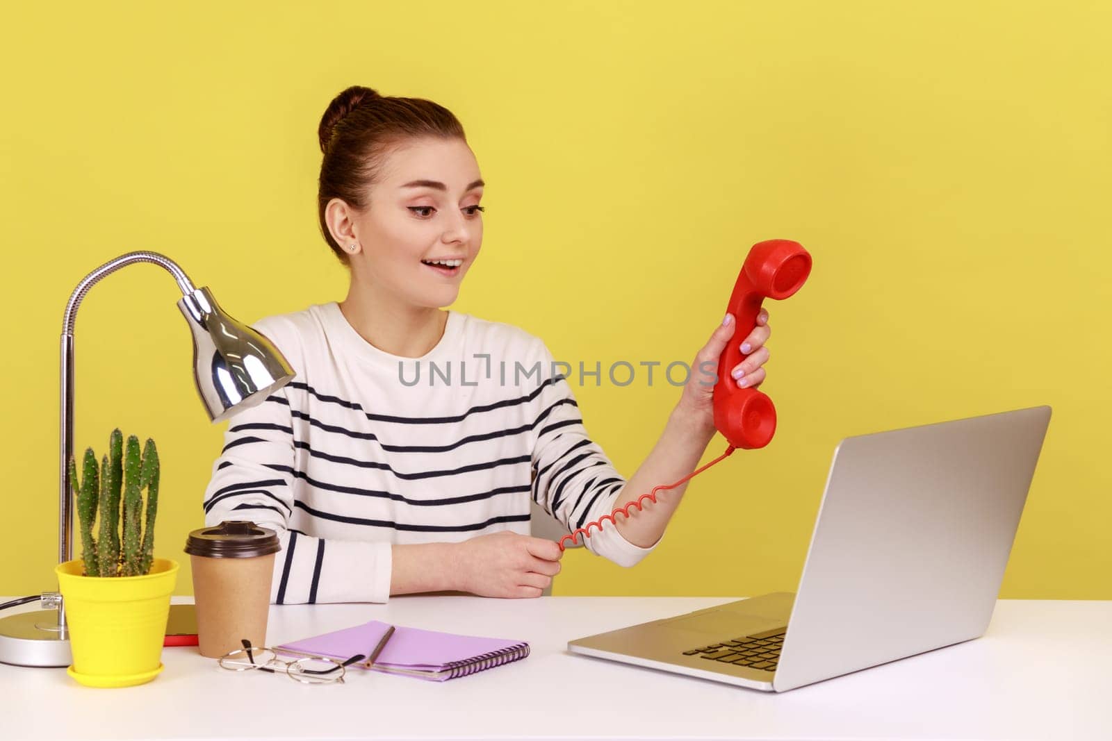 Portrait of smiling happy young adult woman holding and showing retro phone handset to laptop screen, asking to answer phone. Indoor studio studio shot isolated on yellow background.