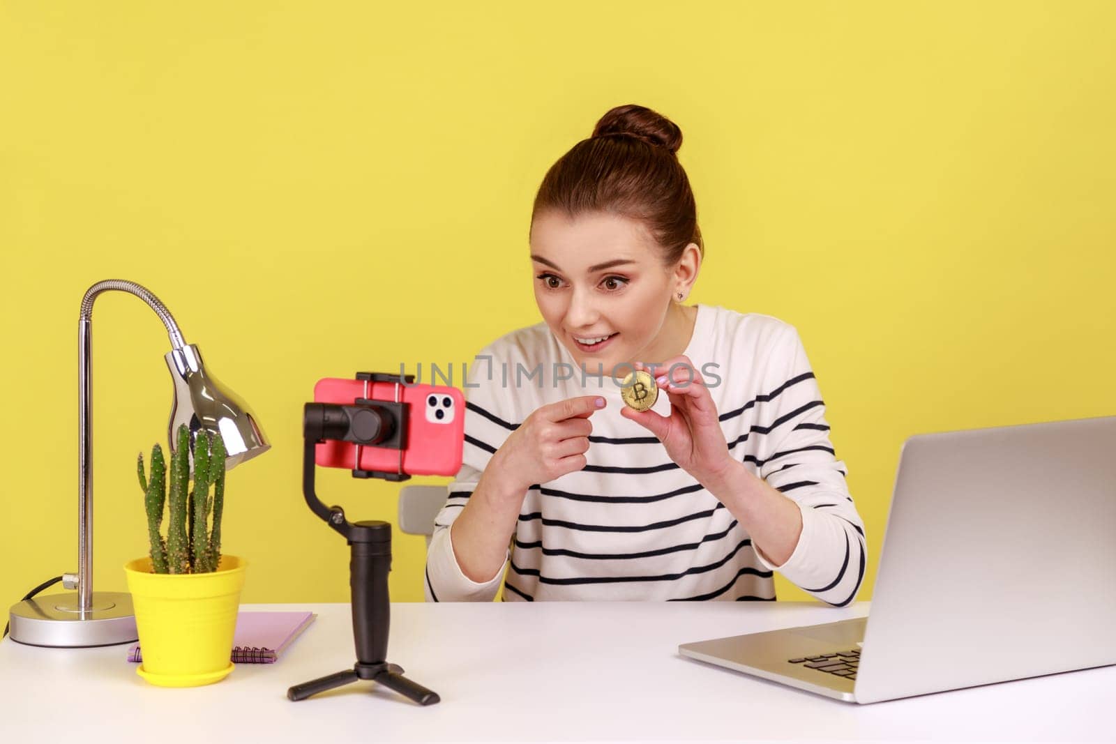 Smiling woman blogger holding in hands and showing on smartphone camera golden bitcoin, explaining how to invest in cryptocurrency, mining. Indoor studio studio shot isolated on yellow background.