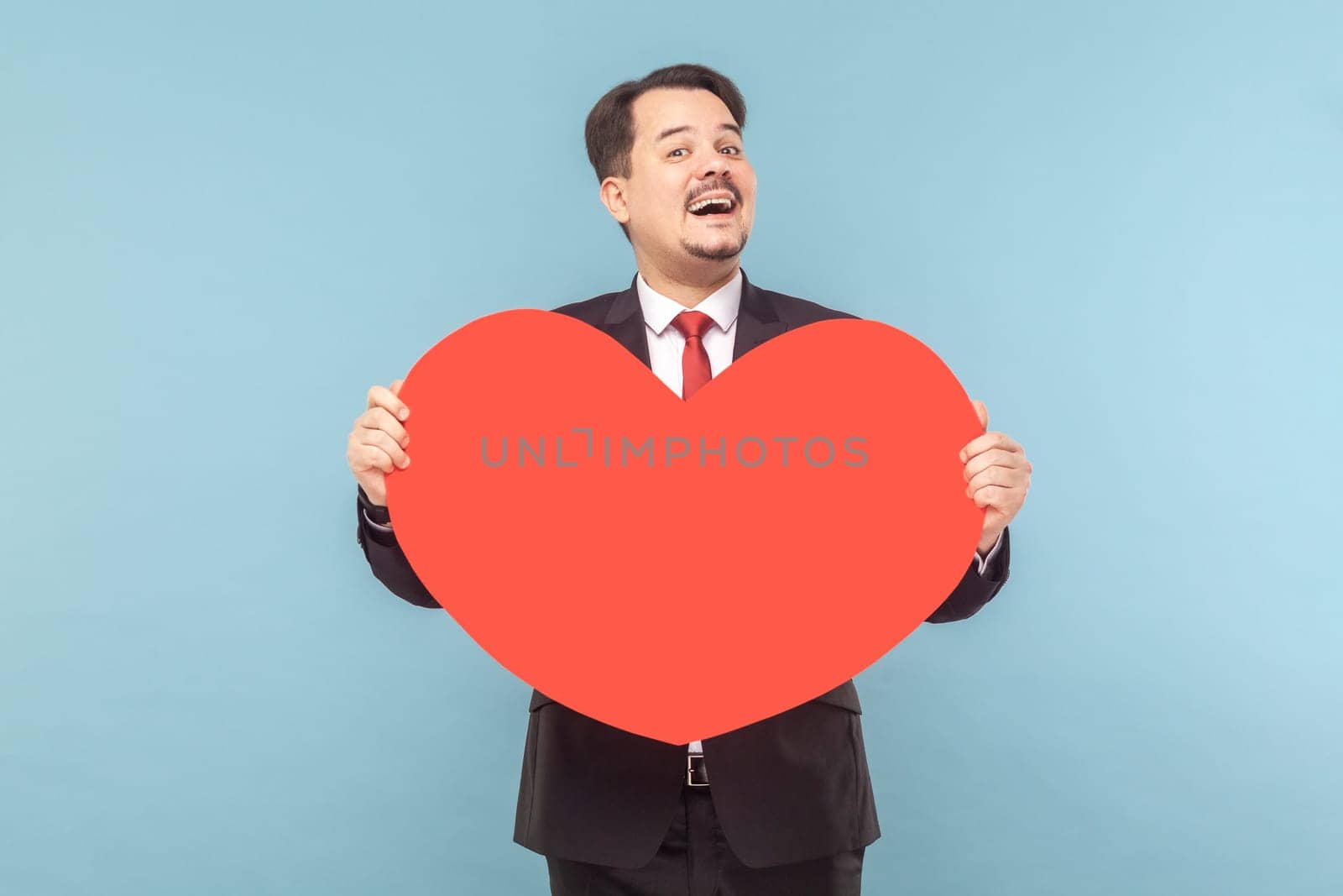 Portrait of satisfied joyful man with mustache standing holding big red card, expressing love and gentle, wearing black suit with red tie. Indoor studio shot isolated on light blue background.