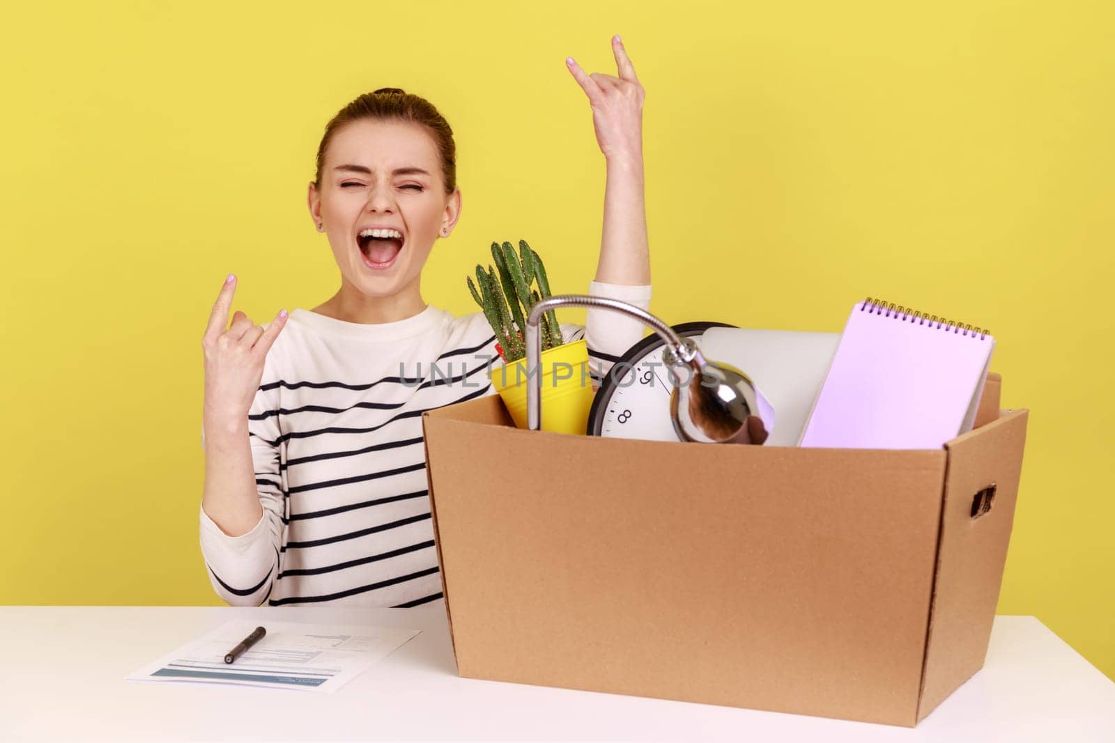 Optimistic woman office worker sitting at workplace with cardboard box with her things and yelling, showing victory sign, rejoice a new job. Indoor studio studio shot isolated on yellow background.