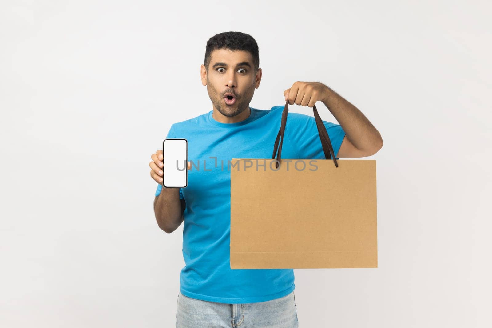 Portrait of shocked astonished handsome man wearing blue T- shirt standing holding shopping bag and mobile phone with blank screen for advertisement. Indoor studio shot isolated on gray background.