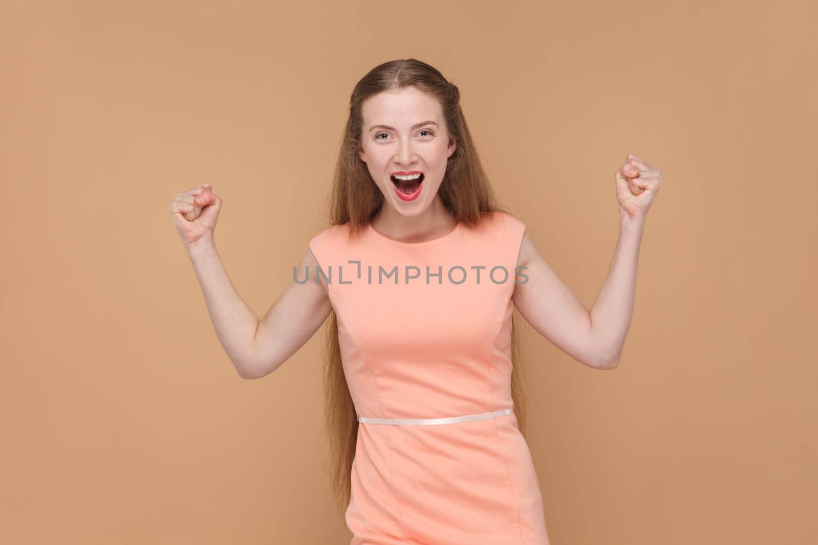 Portrait of extremely happy positive woman with long hair standing with clenched fists, screaming with excitement, wearing elegant dress. Indoor studio shot isolated on brown background.