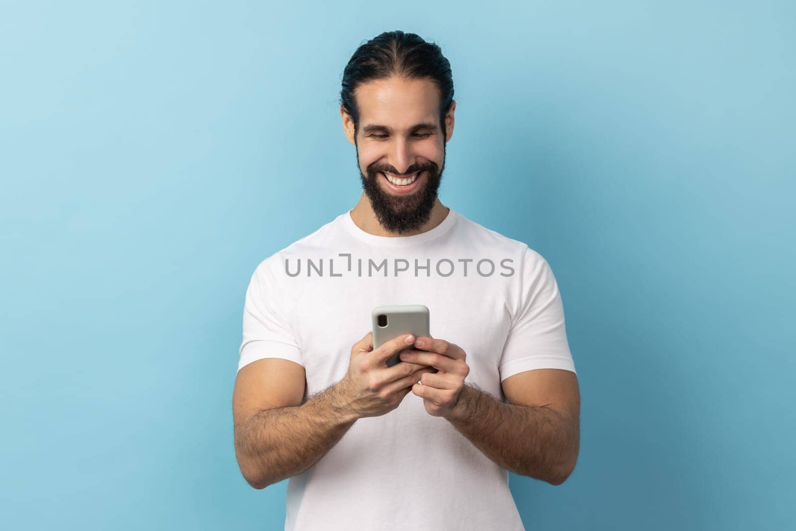 Portrait of handsome positive man bodybuilder with beard wearing white T-shirt using smart phone, looking at device screen, writing post. Indoor studio shot isolated on blue background.