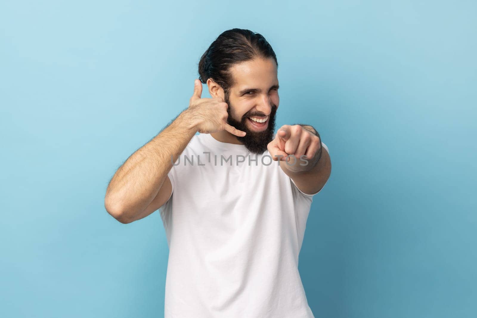 Hey you, call me. Man with beard wearing white T-shirt doing talking on telephone gesture and smiling, flirting, asking contact, pointing to camera. Indoor studio shot isolated on blue background.