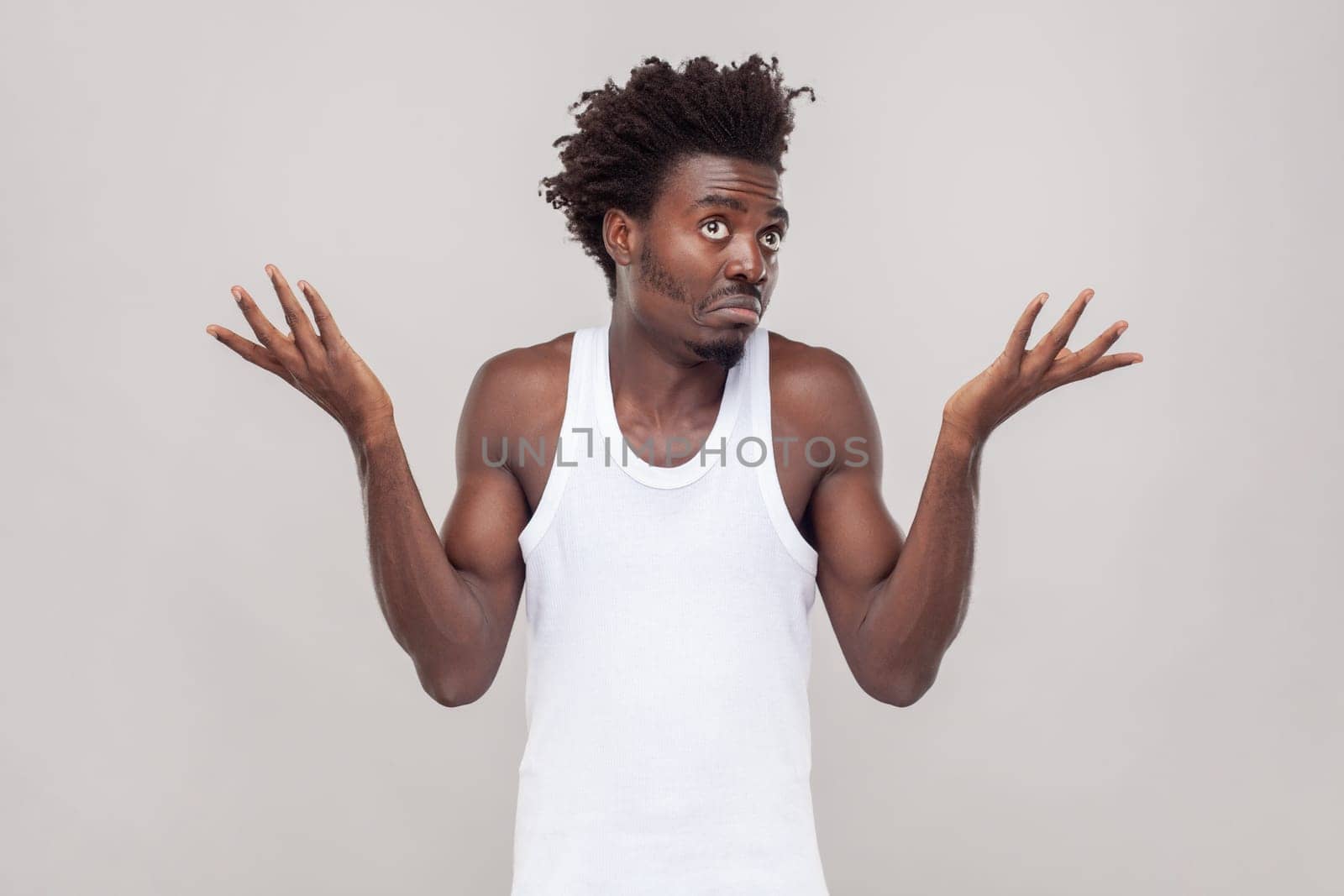 I dont know this. Portrait of confused puzzled man with Afro hairstyle feels uncertain as cant find right way, wearing white T-shirt. Indoor studio shot isolated on gray background.