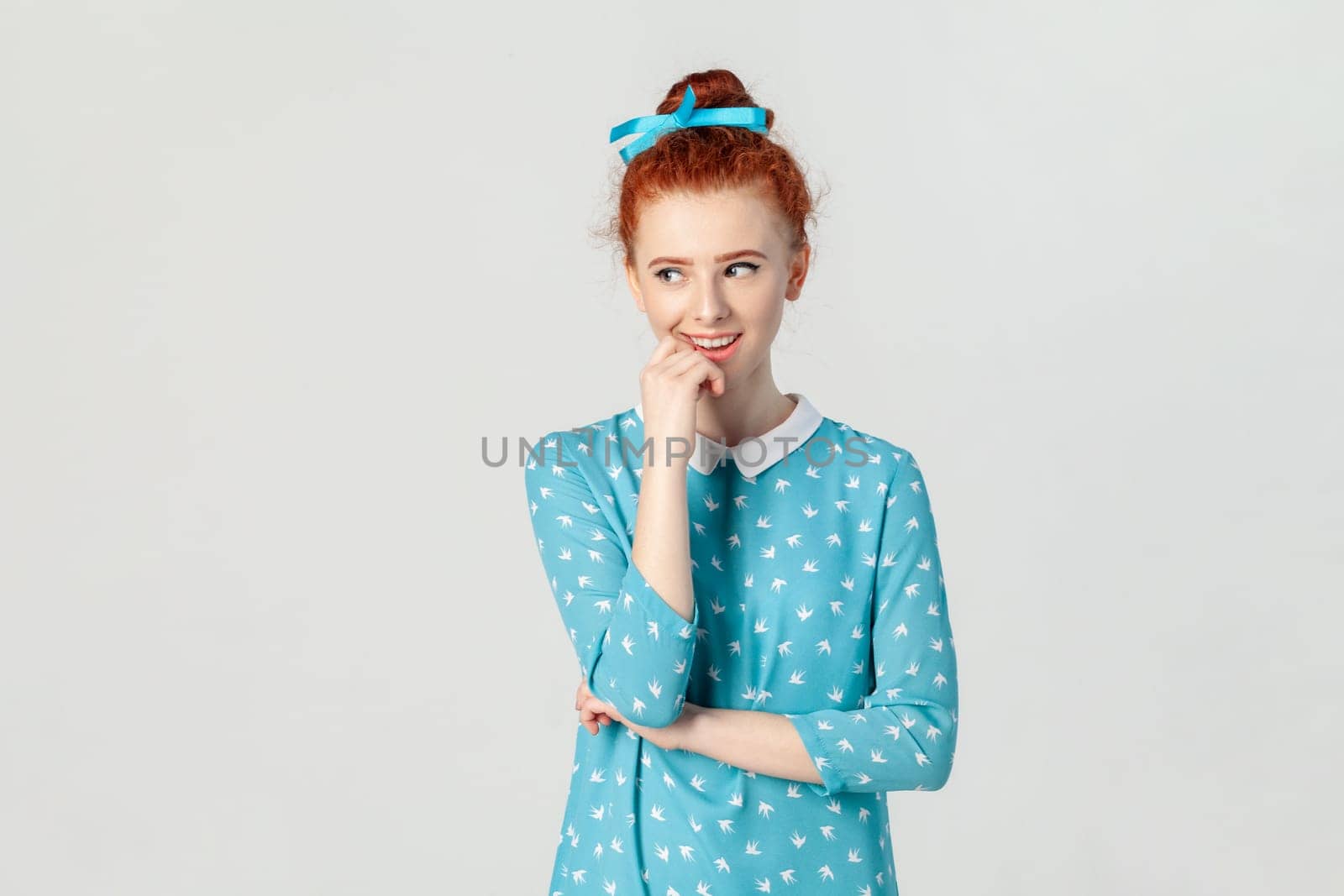 Portrait of sly beautiful red haired woman with hair bun, holding her chin, looking away with cunning expression, planning prank, wearing blue dress. Indoor studio shot isolated on gray background.