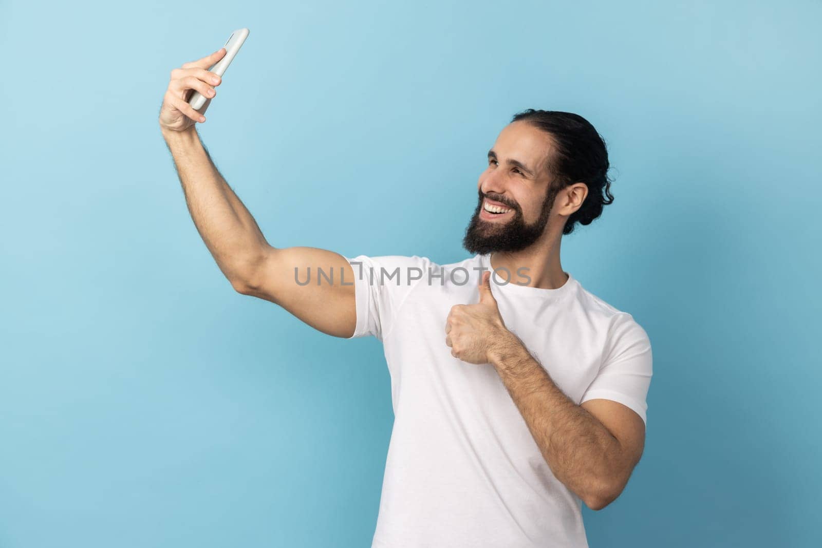 Portrait of man with beard wearing white T-shirt showing thumbs up like gesture and winking looking at phone camera, making selfie or recording video. Indoor studio shot isolated on blue background.