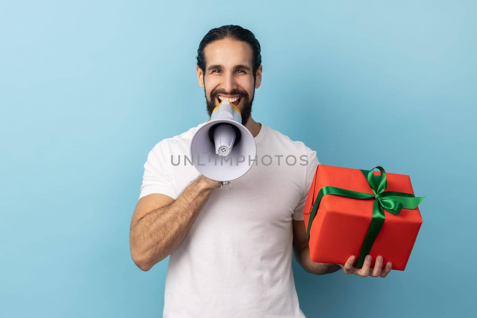 Portrait of positive handsome man with beard wearing white T-shirt holding red present box and screaming in megaphone, making announcement. Indoor studio shot isolated on blue background.