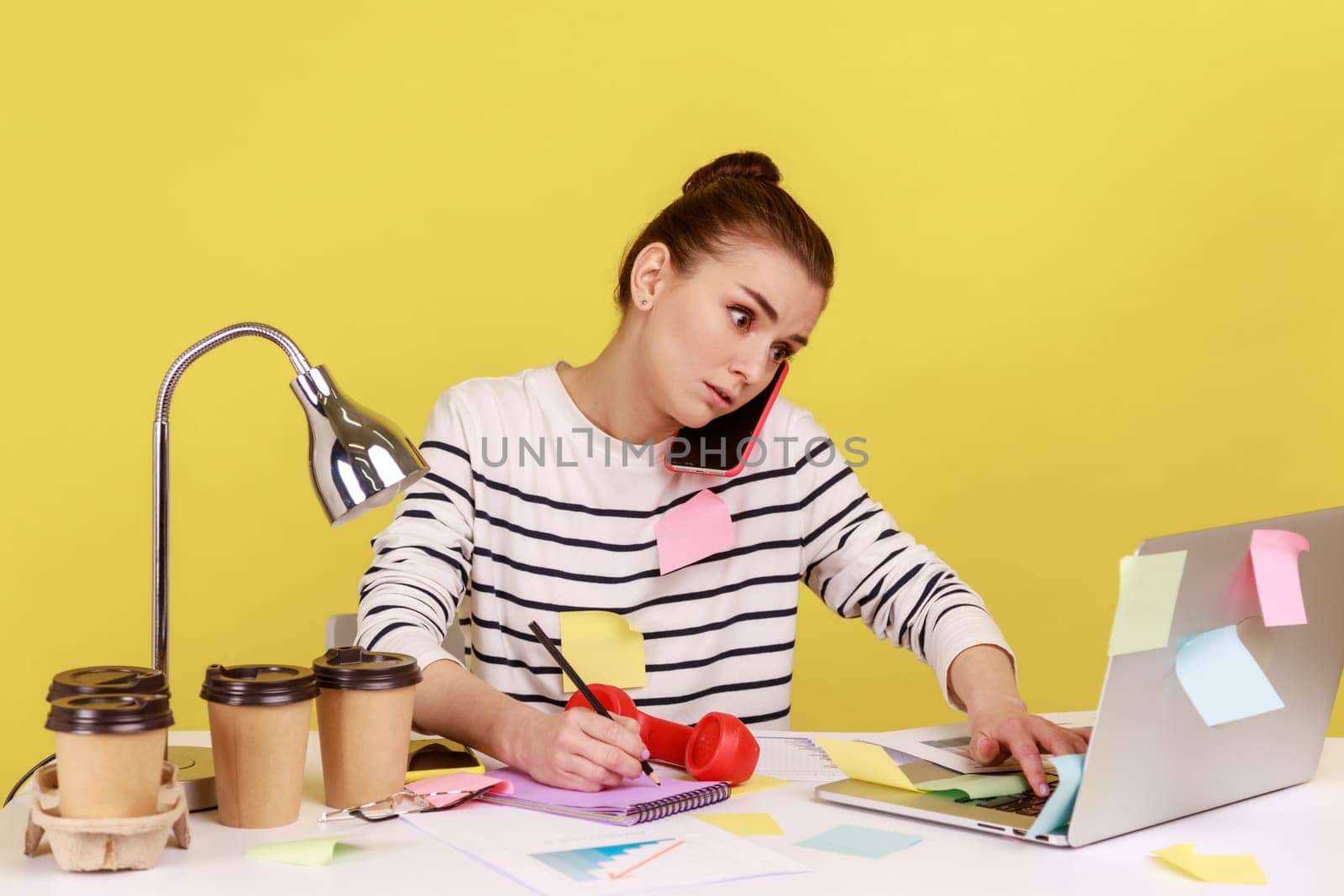 Concentrated secretary in striped shirt receiving calls on phone and making notes, writing down information, sitting at workplace with laptop. Indoor studio studio shot isolated on yellow background.
