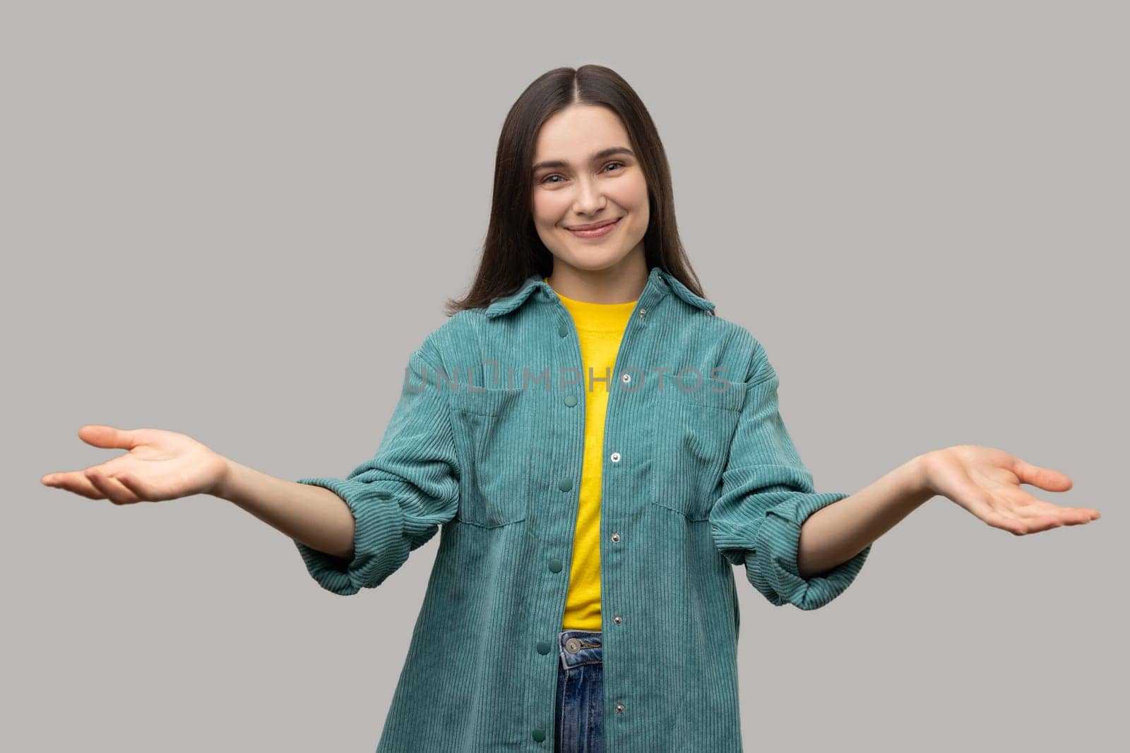 Wide open hug. Nice to meet you. Generous woman standing with wide raised arms and welcoming or sharing, wearing casual style jacket. Indoor studio shot isolated on gray background.