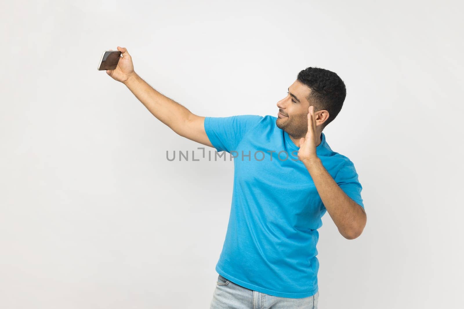 Portrait of friendly positive optimistic man blogger in blue T- shirt holding mobile phone, has video call or livestream, waving hand, hello gesture. Indoor studio shot isolated on gray background.