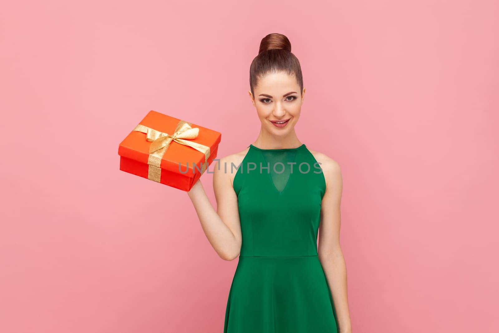 Portrait of attractive positive smiling woman standing with red present box, looking at camera, celebrating holiday, wearing green dress. Indoor studio shot isolated on pink background.
