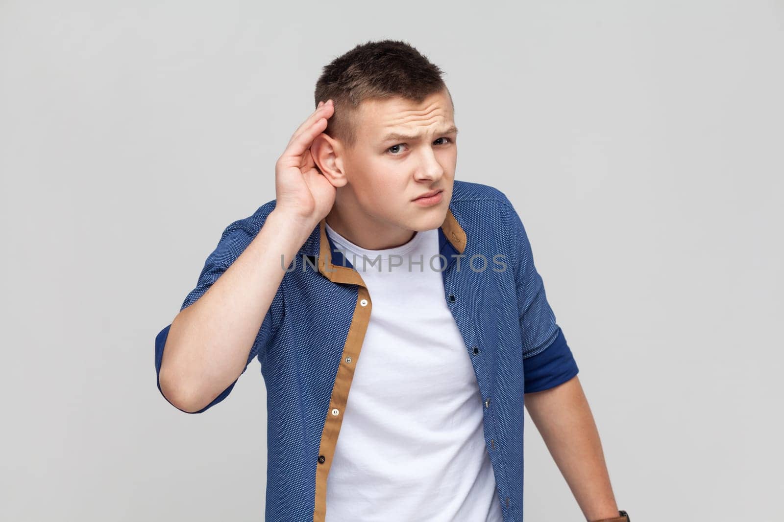 Portrait of teenager boy wearing blue shirt holding hand near ear and listening carefully, having hearing problems, deafness in communication. Indoor studio shot isolated on gray background.