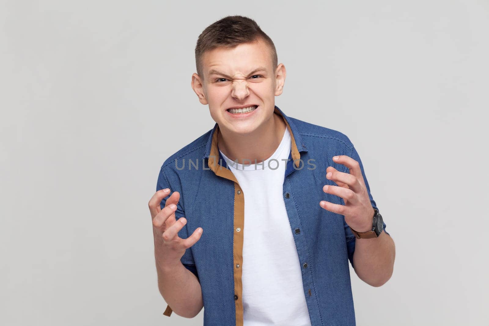 Angry furious teenager boy wearing blue shirt standing and screaming with anger, expressing aggression, swearing with someone, having problems. Indoor studio shot isolated on gray background.