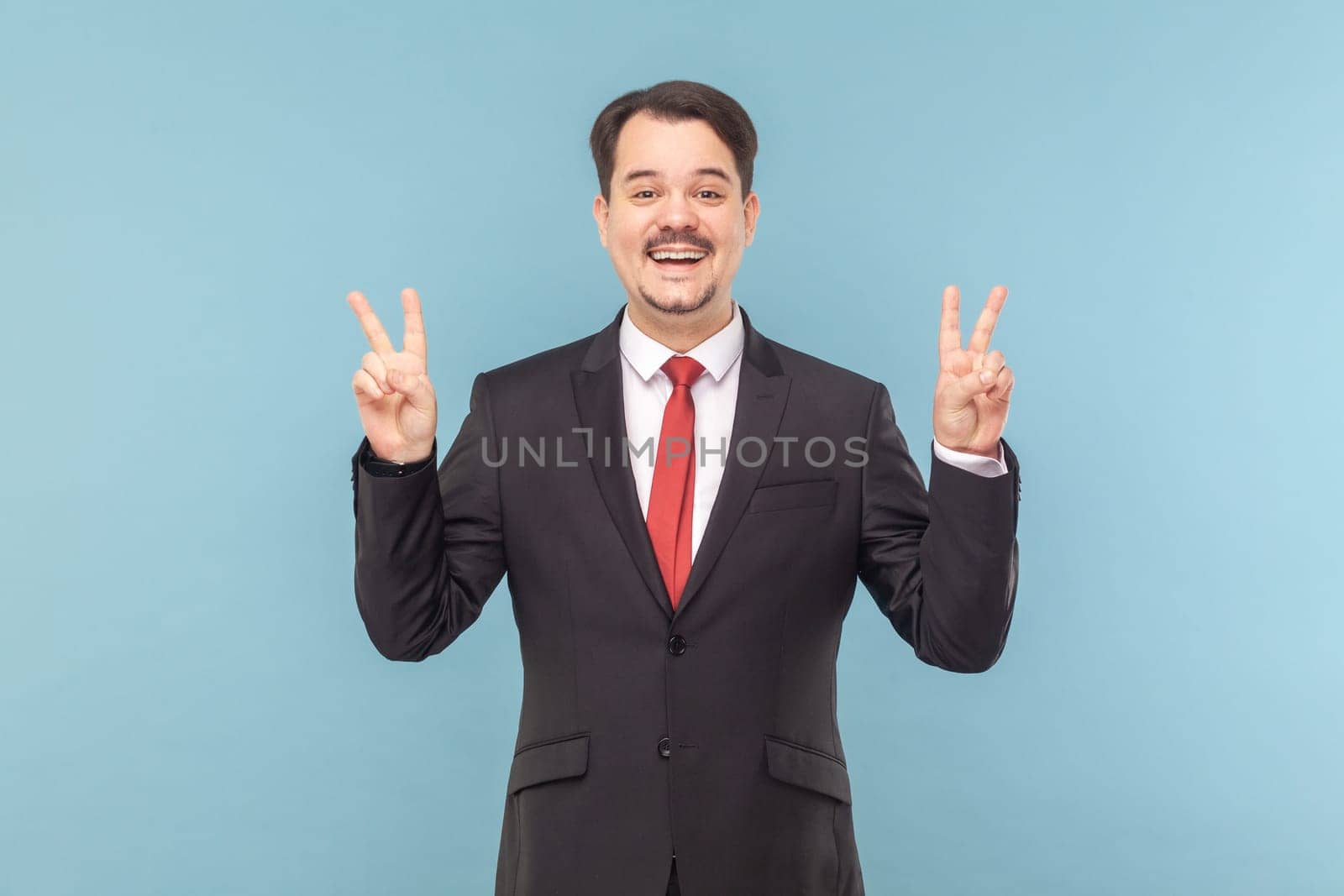 Excited happy positive man with mustache standing and showing v sign, looking at camera with toothy smile, wearing black suit with red tie. Indoor studio shot isolated on light blue background.