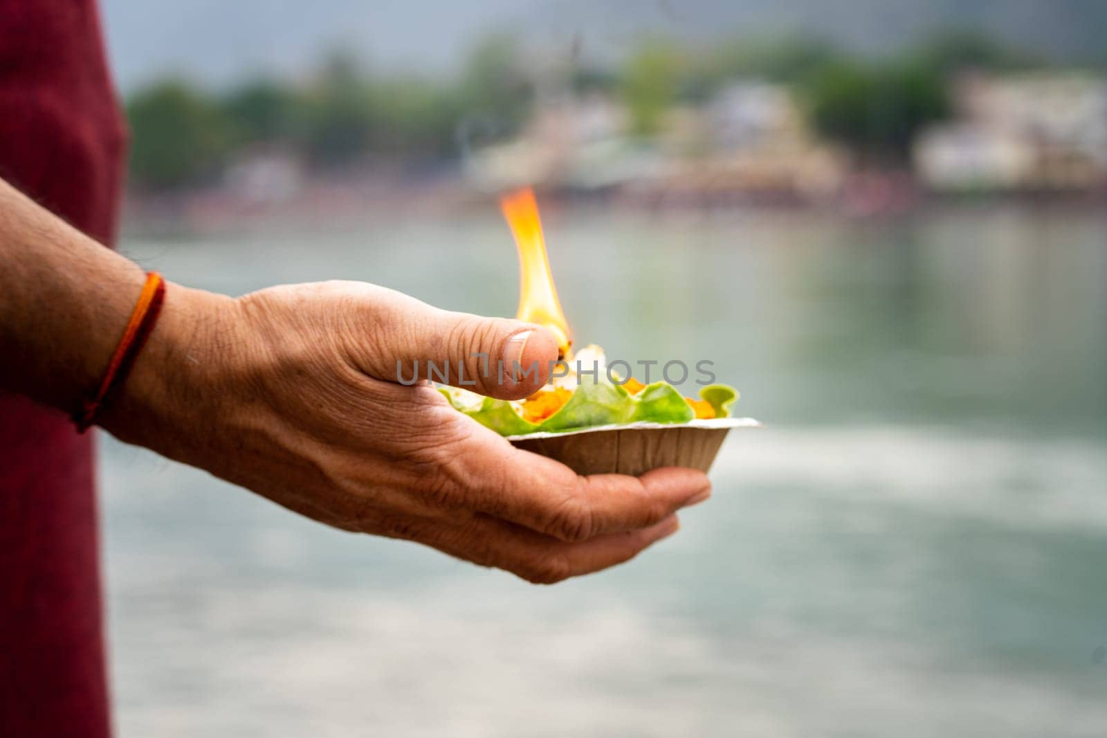 shallow depth of feild shot of man holding leaf plate cup with marigold gande flowers rose and a lit oil ghee lamp diya as an offering to the river ganga at rishikesh by Shalinimathur
