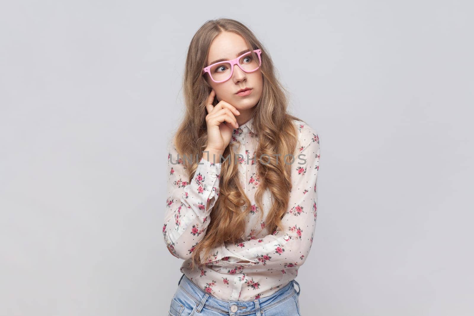 Let me think. Portrait of woman in glasses with wavy blond hair standing with puzzled serious expression, thinking doubting, making choice. Indoor studio shot isolated on gray background.