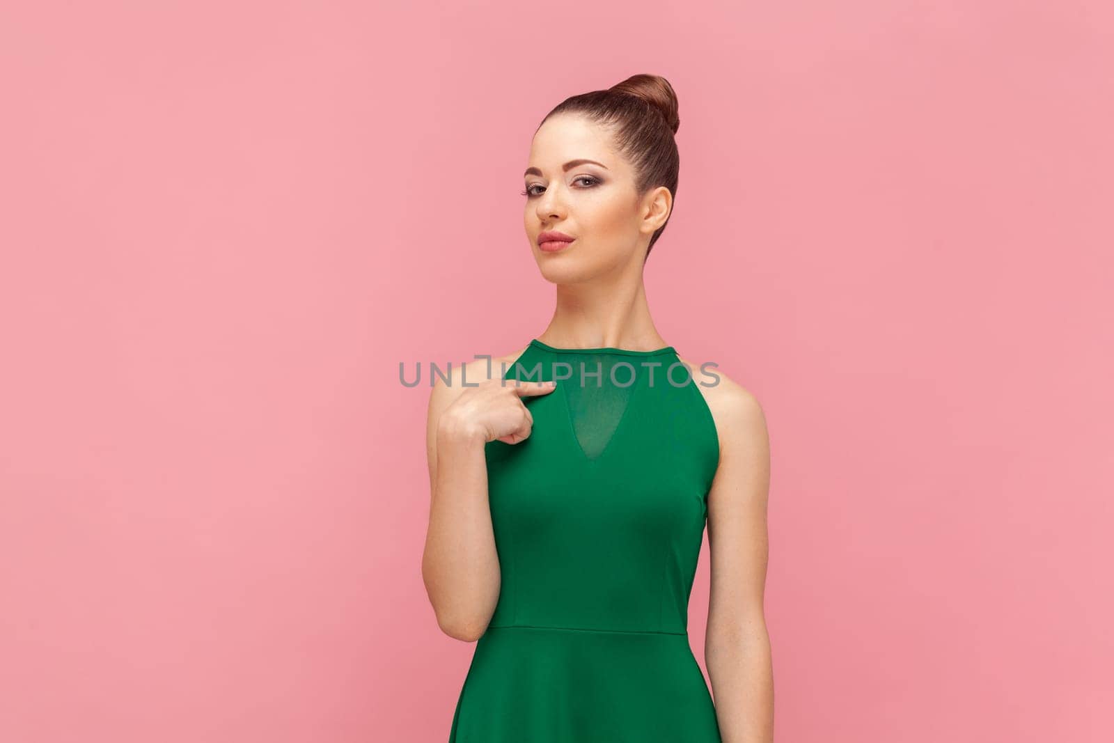Portrait of self-confident egoistic woman with bun hairstyle standing with proud facial expression, pointing finger at herself , wearing green dress. Indoor studio shot isolated on pink background.