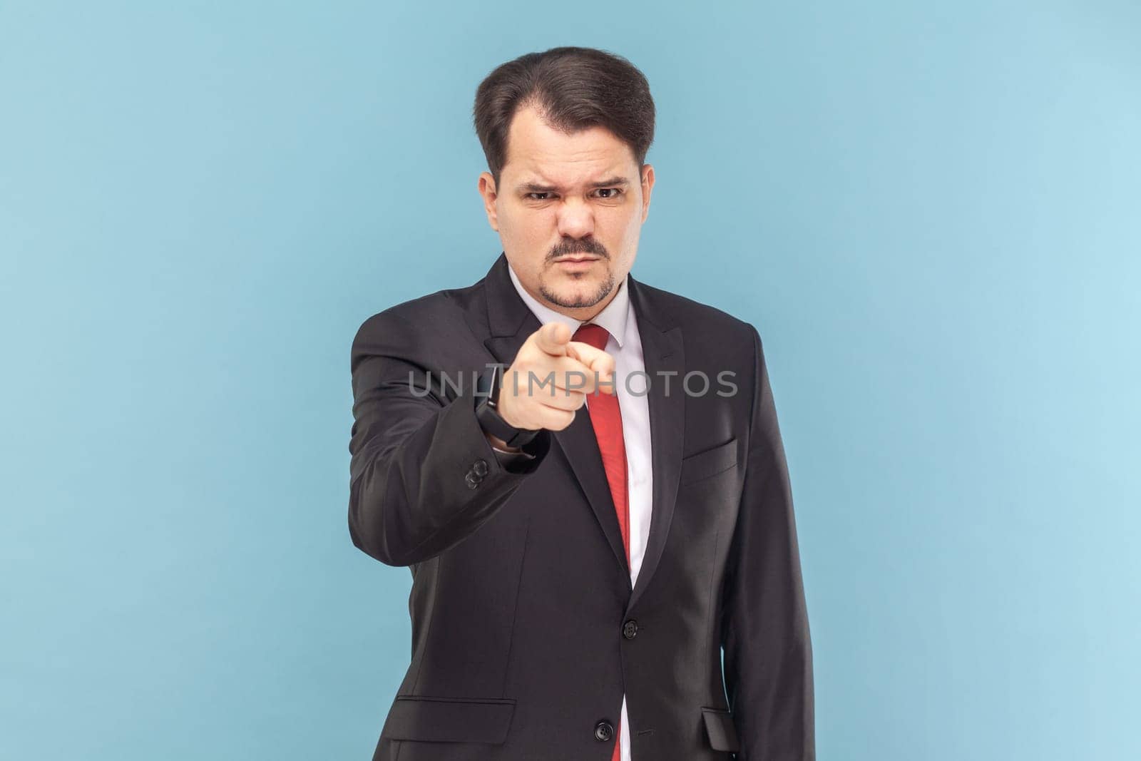 Portrait of serious strict bossy man standing indicating finger to camera, selecting you, looking with anger, wearing black suit with red tie. Indoor studio shot isolated on light blue background.