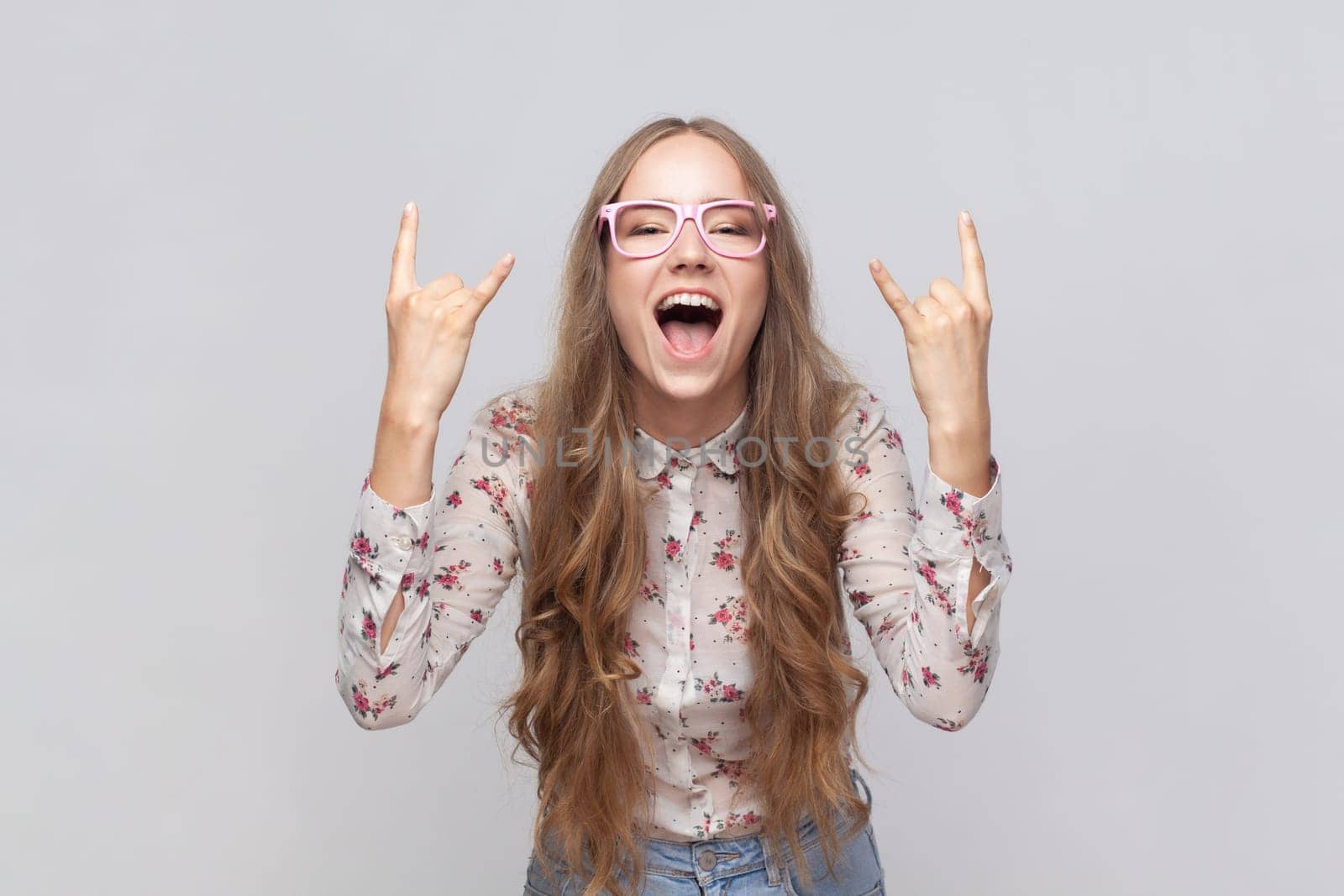 Rock n roll. Portrait of woman in glasses with wavy blond hair standing with rock sing, screaming yelling with excited facial expression. Indoor studio shot isolated on gray background.