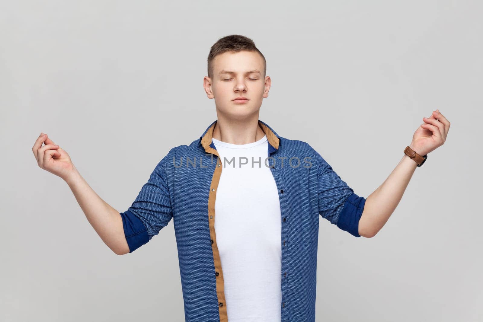 Portrait of calm meditative handsome teenager boy wearing blue shirt standing with closed eyes and yoga gesture pose, trying to calm down. Indoor studio shot isolated on gray background.