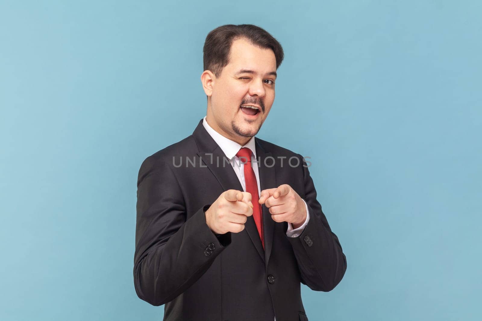 Portrait of flirting positive optimistic man with mustache standing pointing to camera, choosing you, wearing black suit with red tie. Indoor studio shot isolated on light blue background.