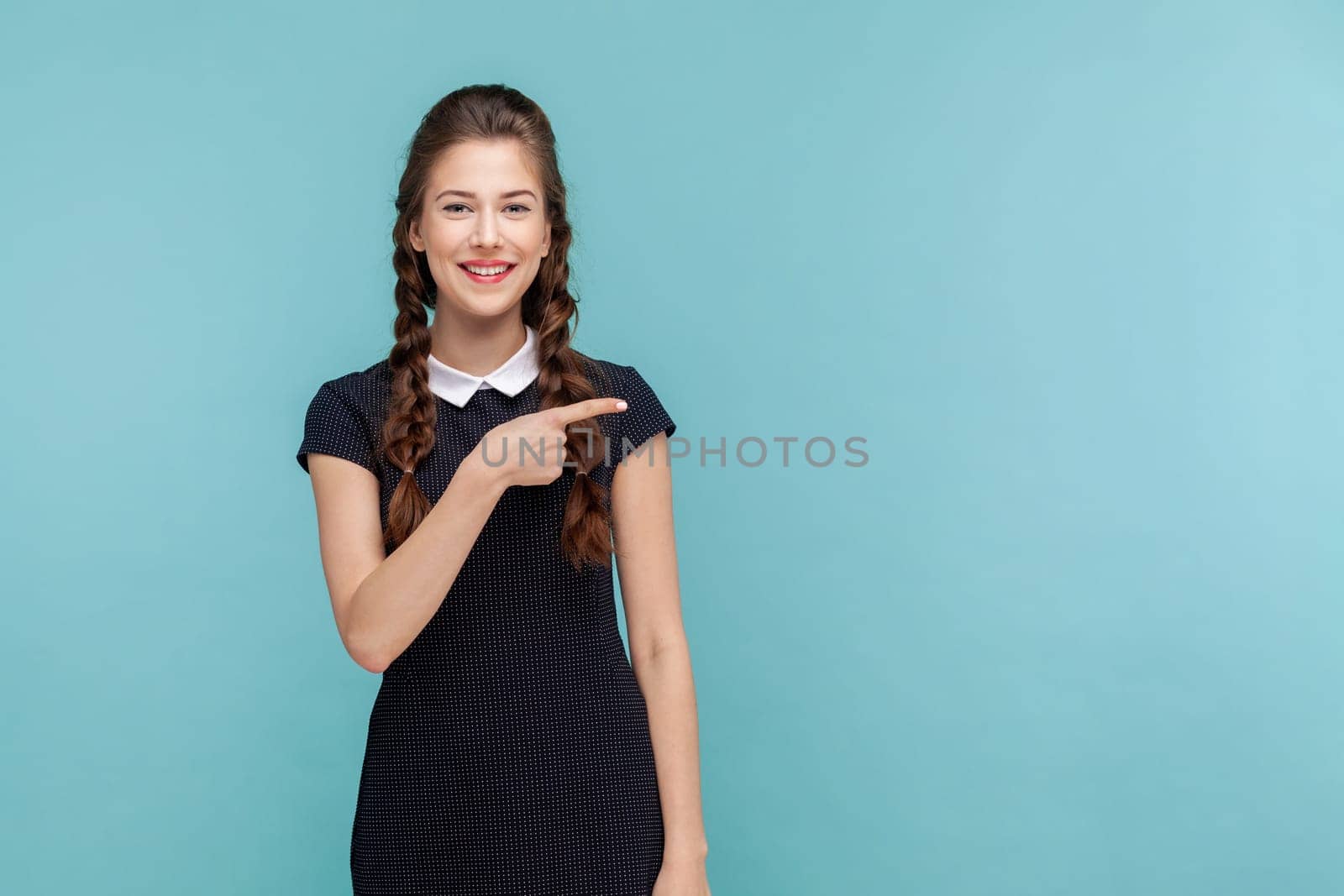 Your advertising here. Portrait of woman points away on copy space, suggests follow this direction or click on link, wearing black dress. woman Indoor studio shot isolated on blue background.