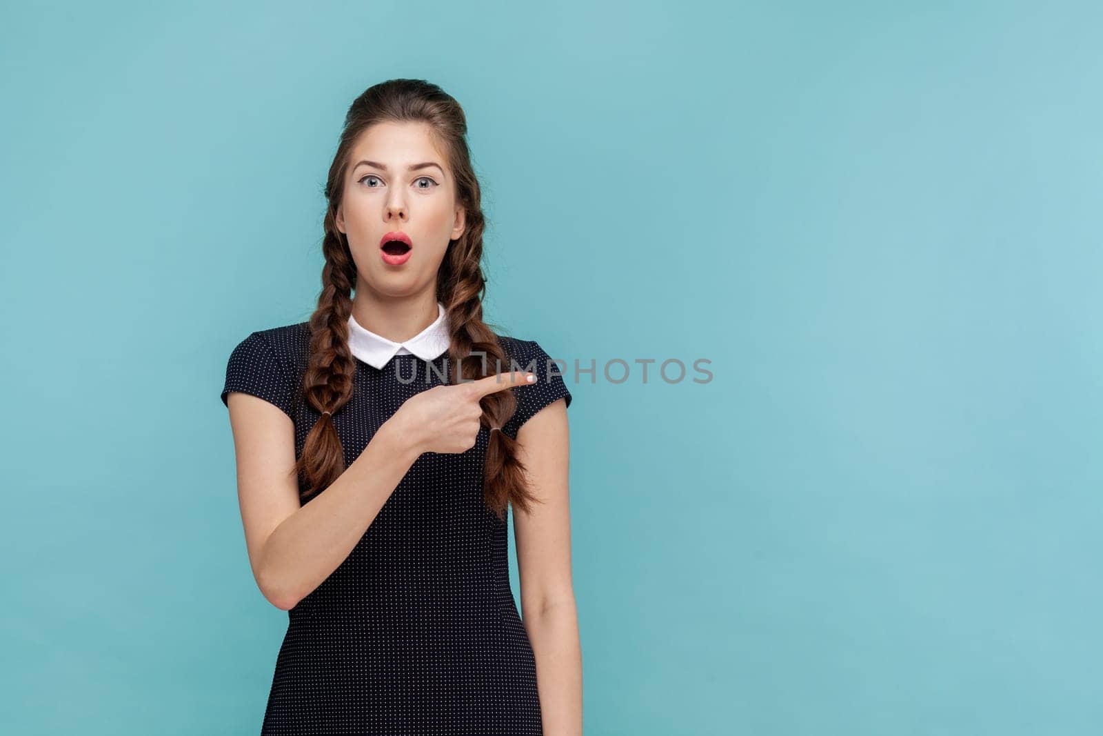 Portrait of shocked surprised woman points away on copy space, suggests follow this direction or click on link, wearing black dress. woman Indoor studio shot isolated on blue background.