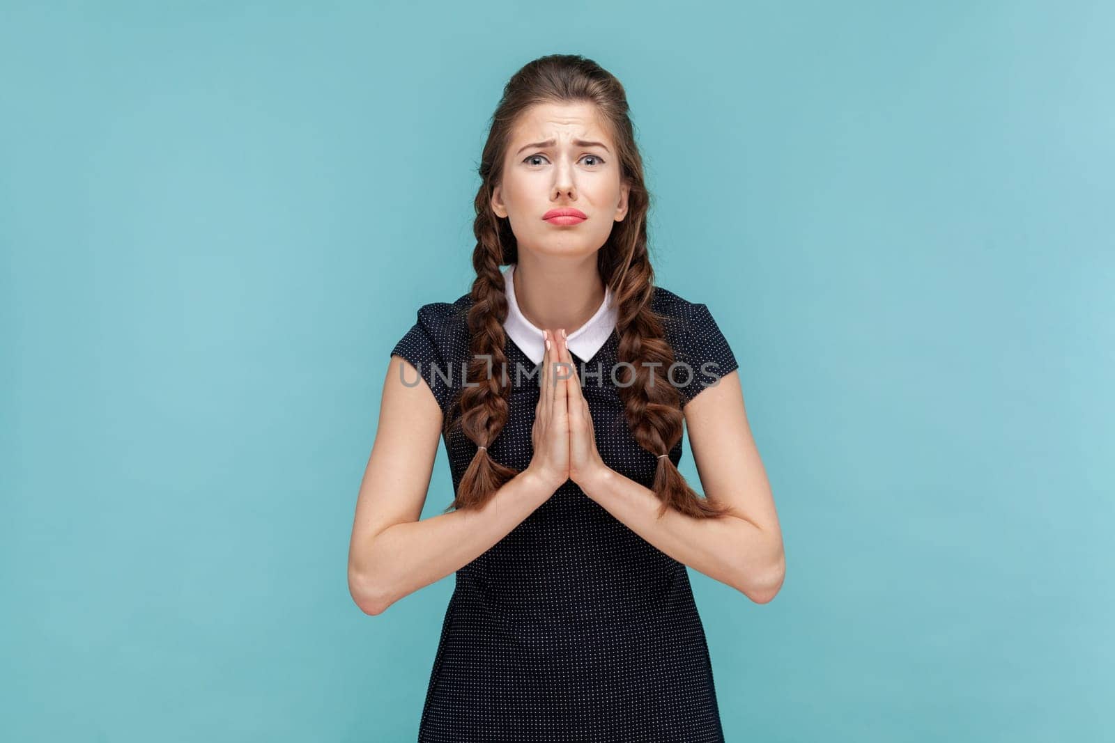 Portrait of hopeful uneasy woman with braids begging for forgiveness keeps palms in praying gesture, asks mercy and says please, needs your help. woman Indoor studio shot isolated on blue background