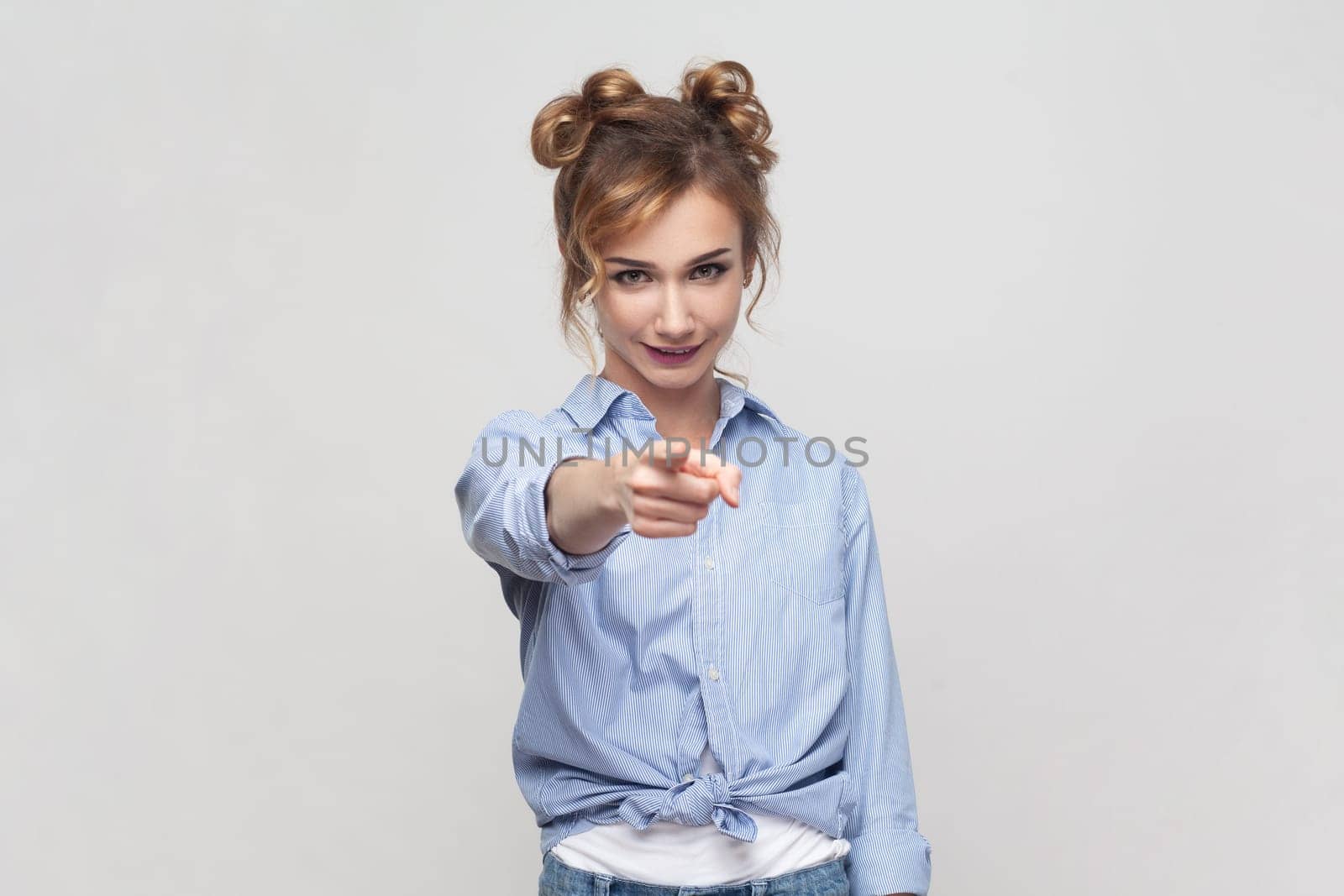 Portrait of joyous pretty blonde woman pointing finger to camera, expresses choice, smiling broadly, saying you are chosen, wearing blue shirt. Indoor studio shot isolated on gray background.