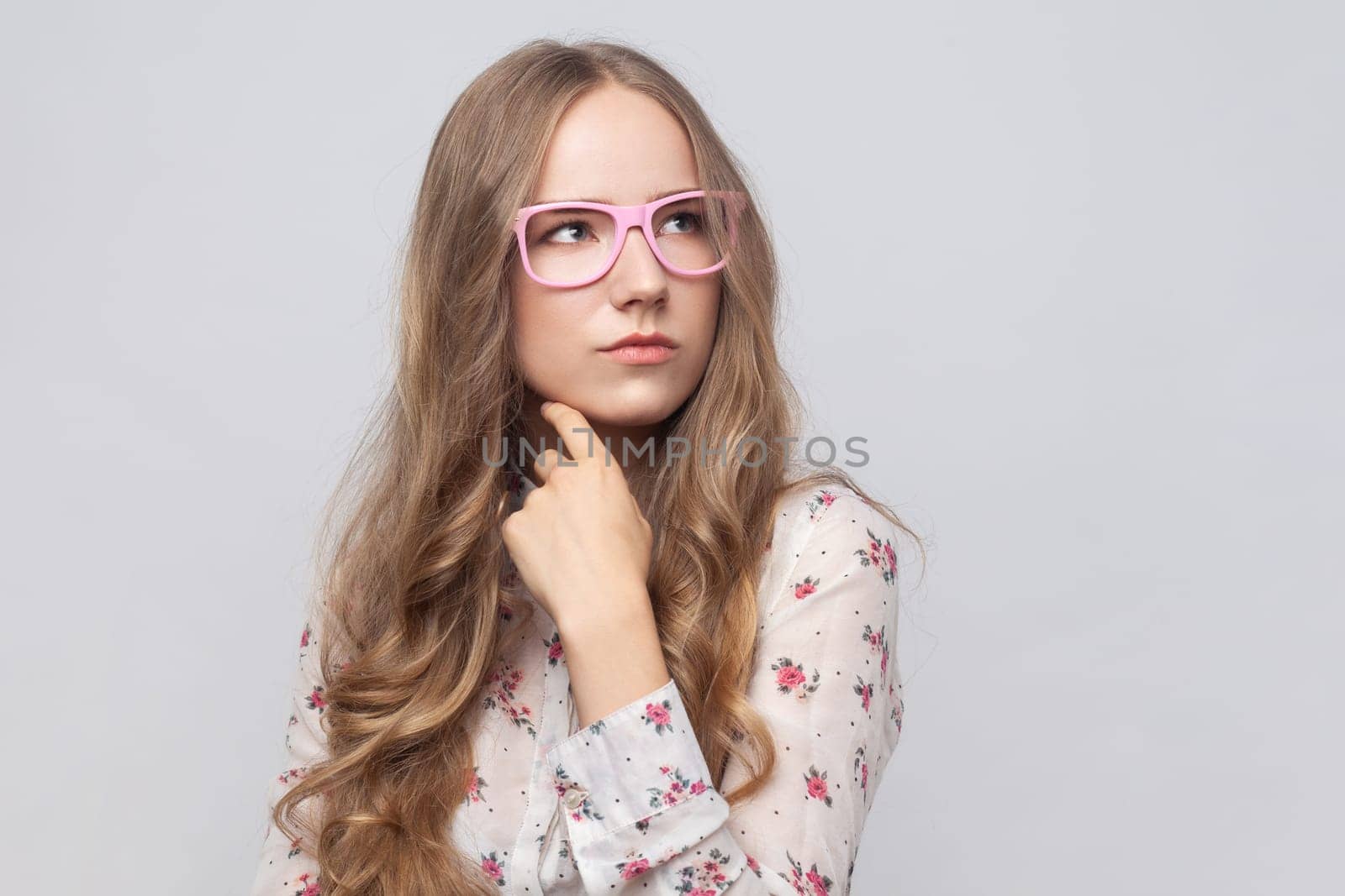 Portrait of thoughtful pensive woman in glasses with long blond hair thinking about future, holding chin, having serious facial expression. Indoor studio shot isolated on gray background.