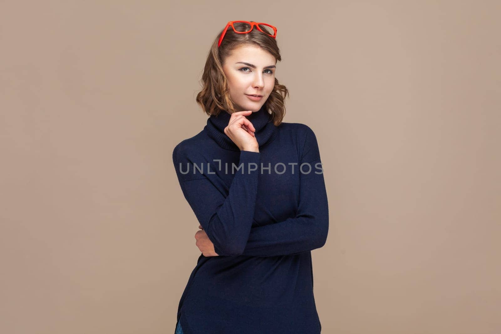 Portrait of self confident attractive woman with wavy hair in red red glasses on her head looking at camera, holding her chin. Indoor studio shot isolated on light brown background.