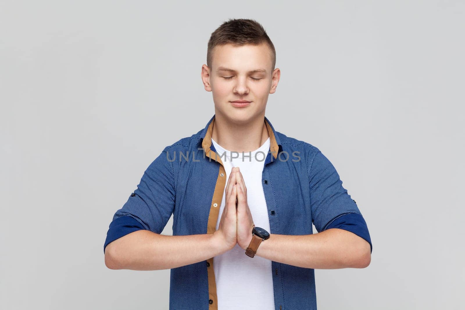 Portrait of calm attractive smiling teenager boy wearing blue shirt standing in yoga pose and try to relaxing, keeps palms together. Indoor studio shot isolated on gray background.