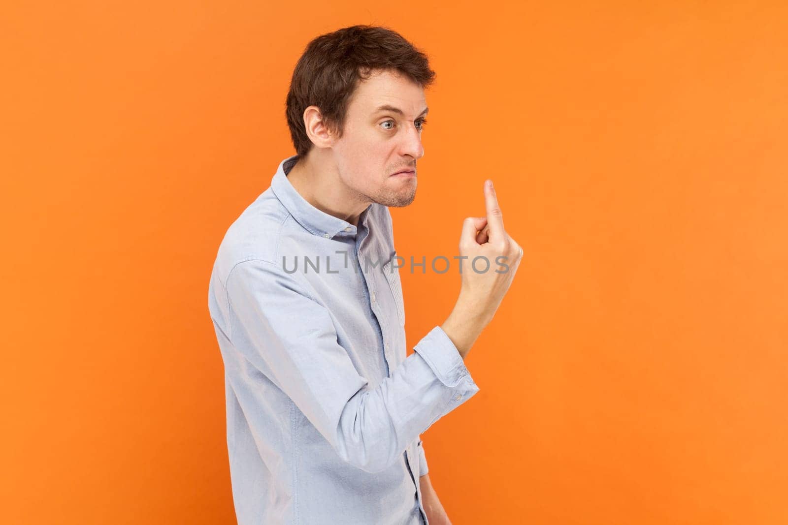 Side view portrait of angry rude man standing looking away with aggression, showing middle finger, fighting with someone, wearing light blue shirt. Indoor studio shot isolated on orange background.