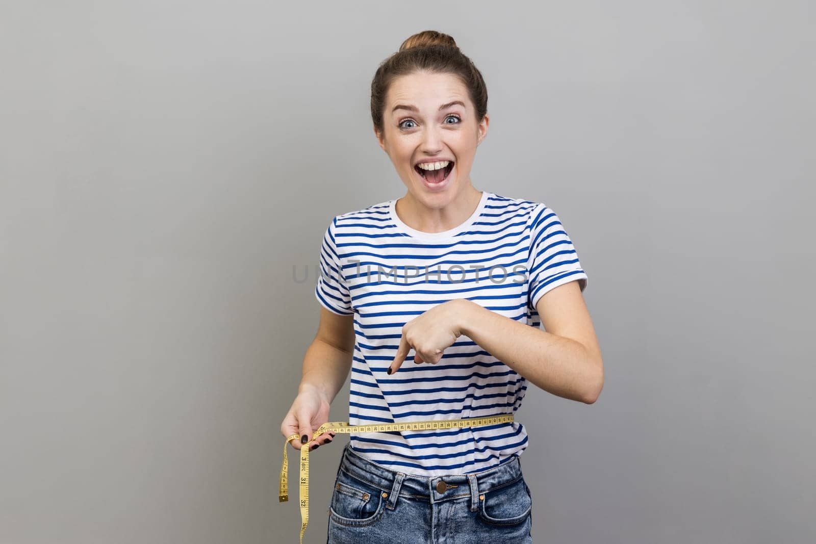 Portrait of woman wearing striped T-shirt holding on waist tape, pointing and looking at camera with satisfaction and excitement. Indoor studio shot isolated on gray background.