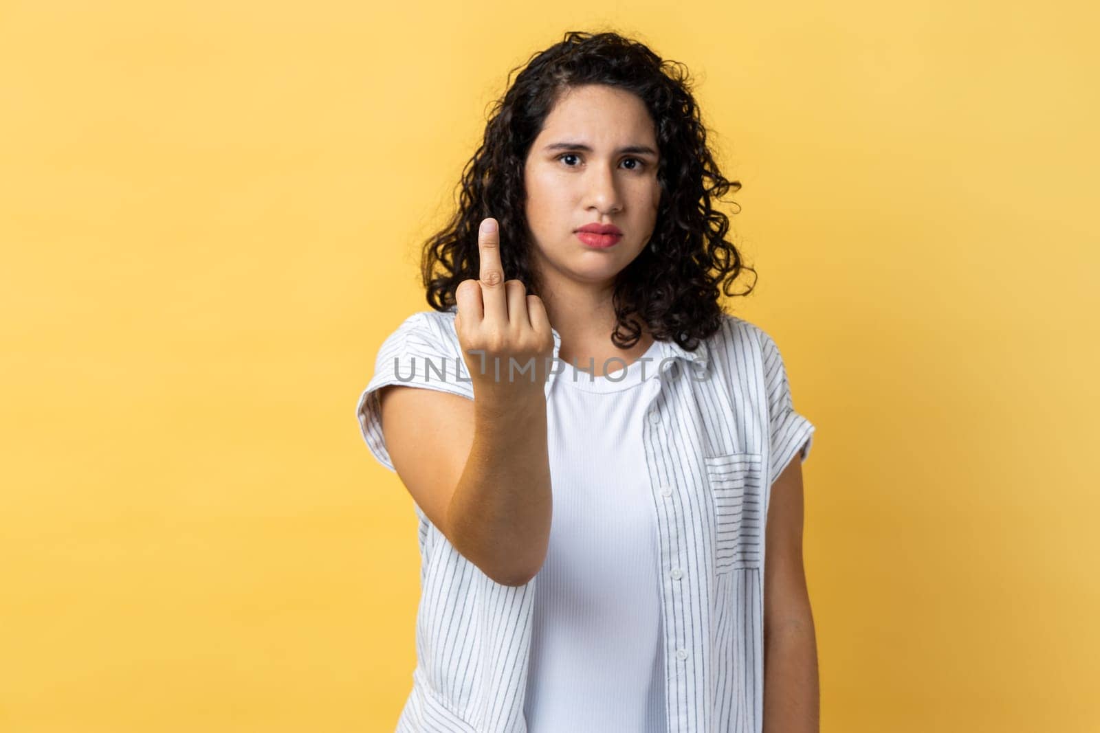 Portrait of woman with dark wavy hair showing middle finger and asking to get off looking at camera with negativity, disrespectful behaviour. Indoor studio shot isolated on yellow background.