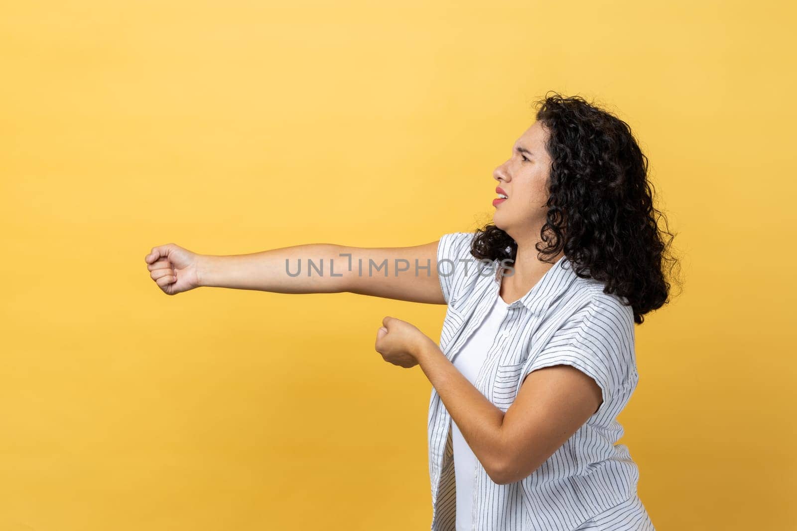 Side view portrait of attractive hard working woman with dark wavy hair standing and doing pulling gesture of something, making effort. Indoor studio shot isolated on yellow background.